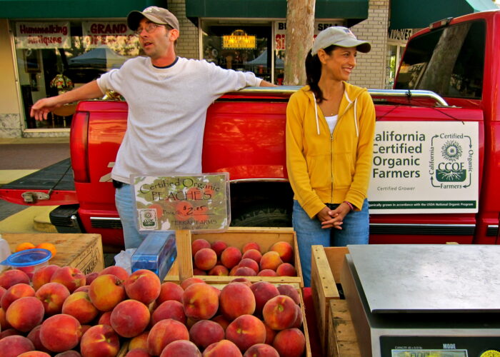Two farmers at a market stand with peaches, promoting California Certified Organic Farmers.