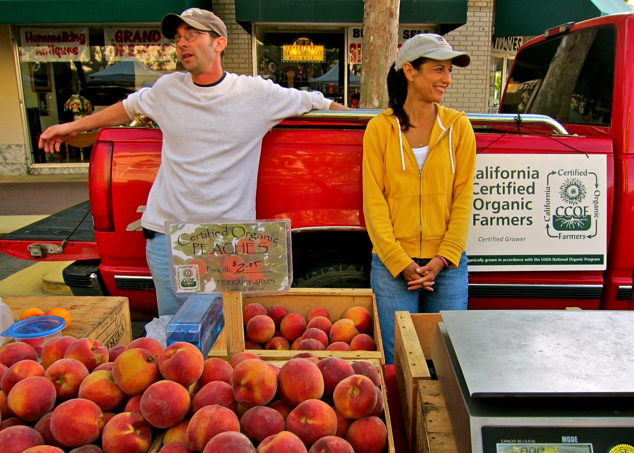 Two farmers at a market stand with peaches, promoting California Certified Organic Farmers.