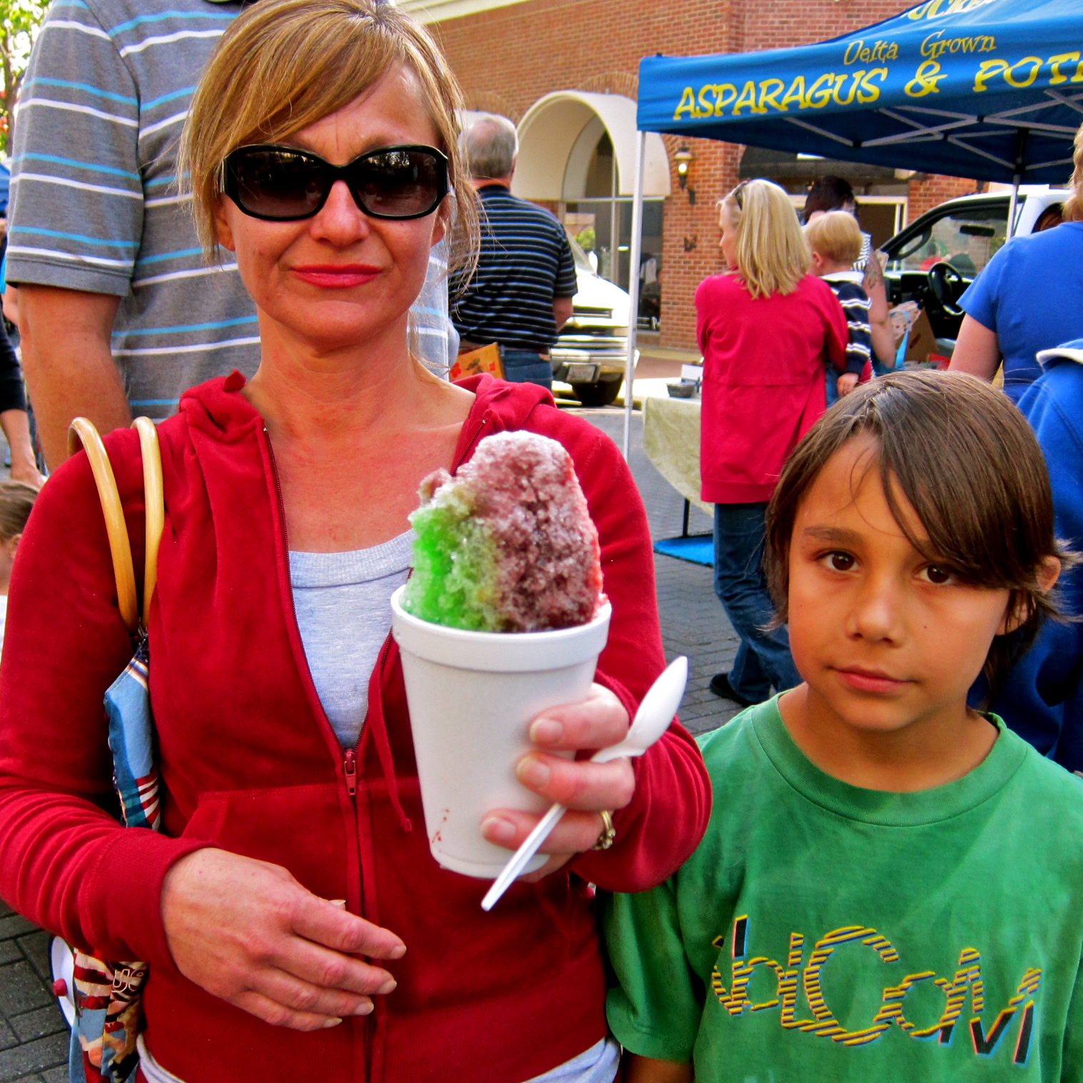 Woman and child at a local event, woman holding colorful shaved ice, outdoor setting with tents and people.