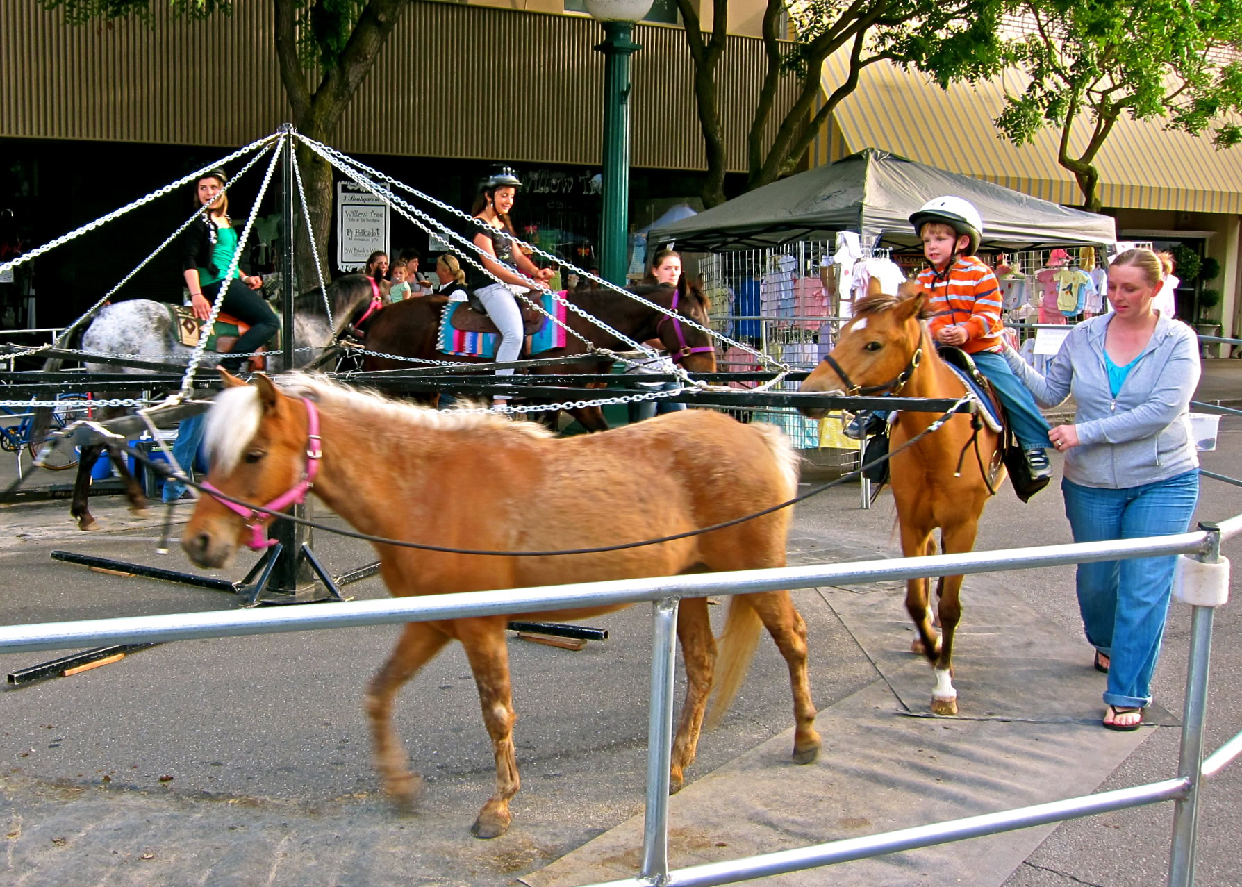 Children riding ponies at a fair with onlookers and a tent in the background.