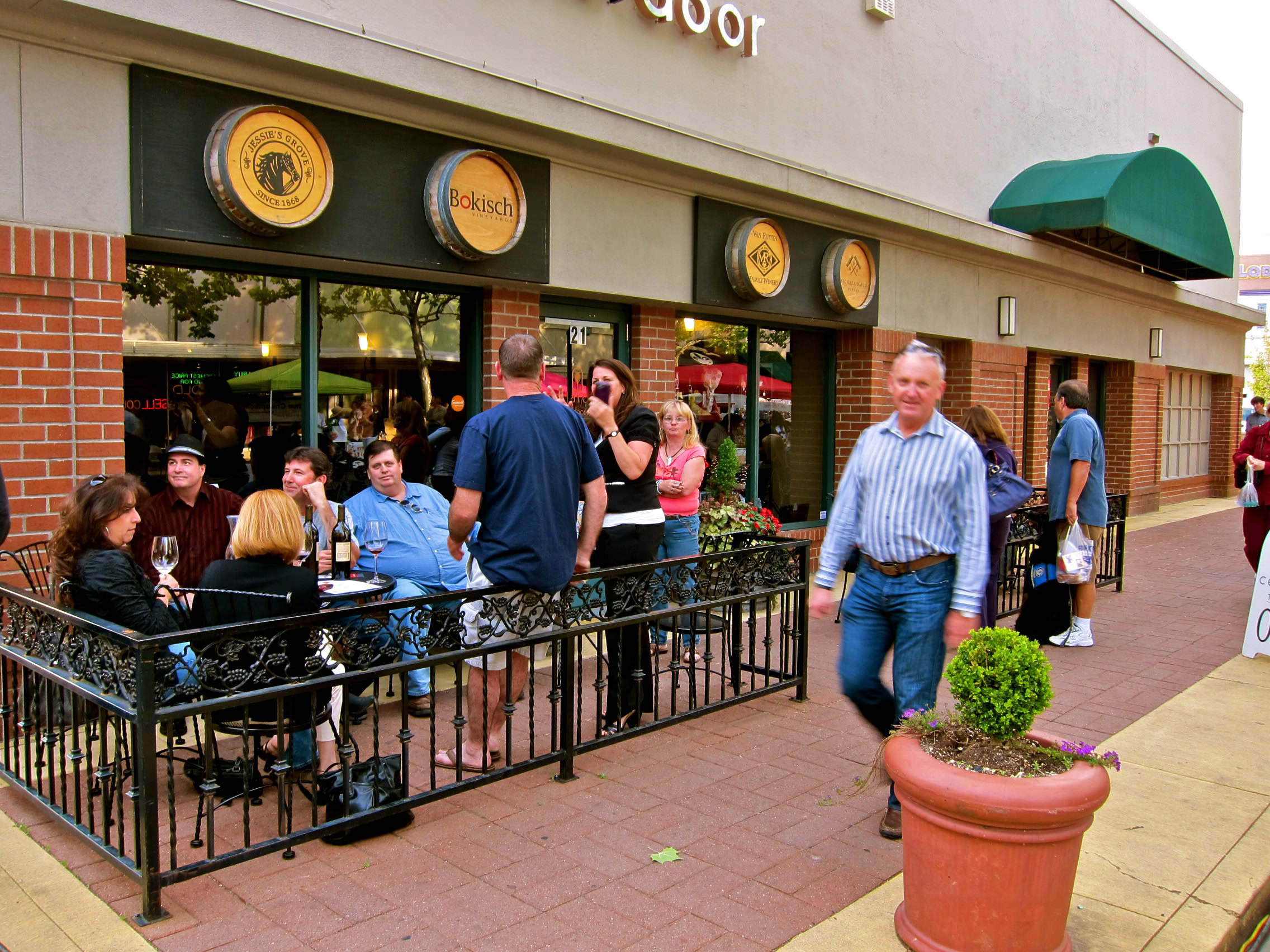 People enjoying outdoor dining at a Lodi wine bar with wine barrels decor and a green awning.