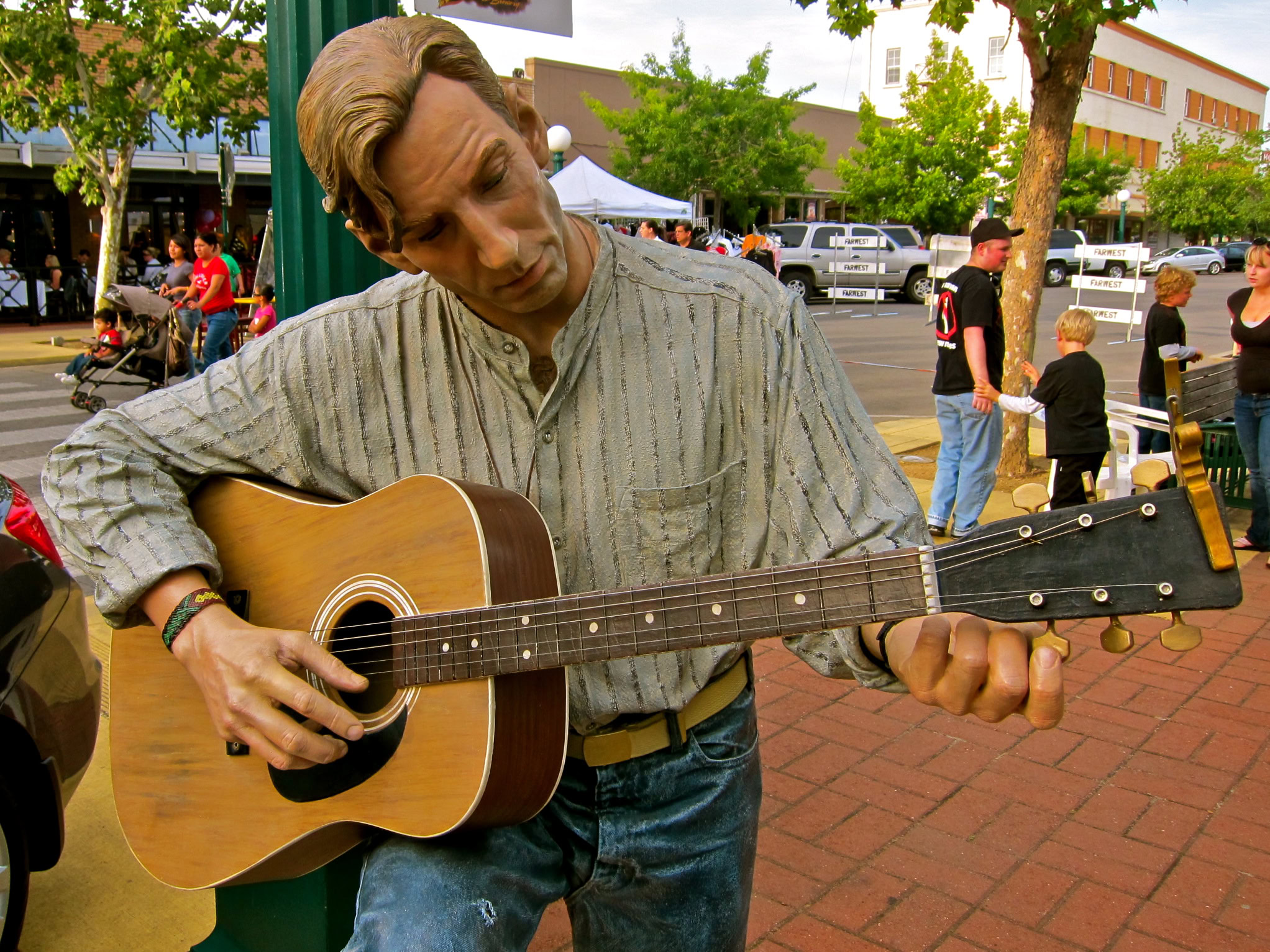 Male street musician playing acoustic guitar at outdoor event with crowd and trees in background.
