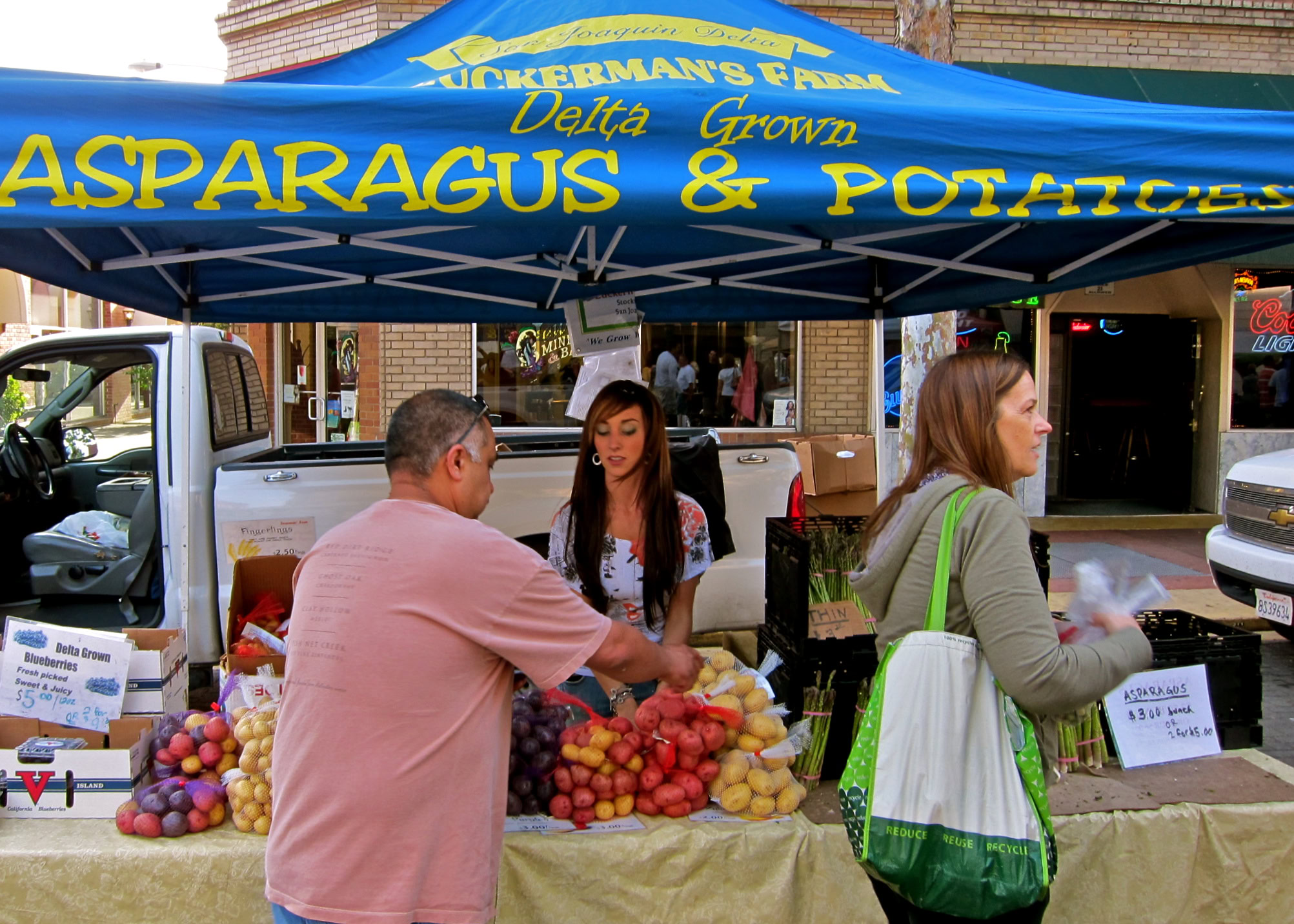 People shopping at a farmers market stall with fresh asparagus, potatoes, and grapes under a blue canopy.