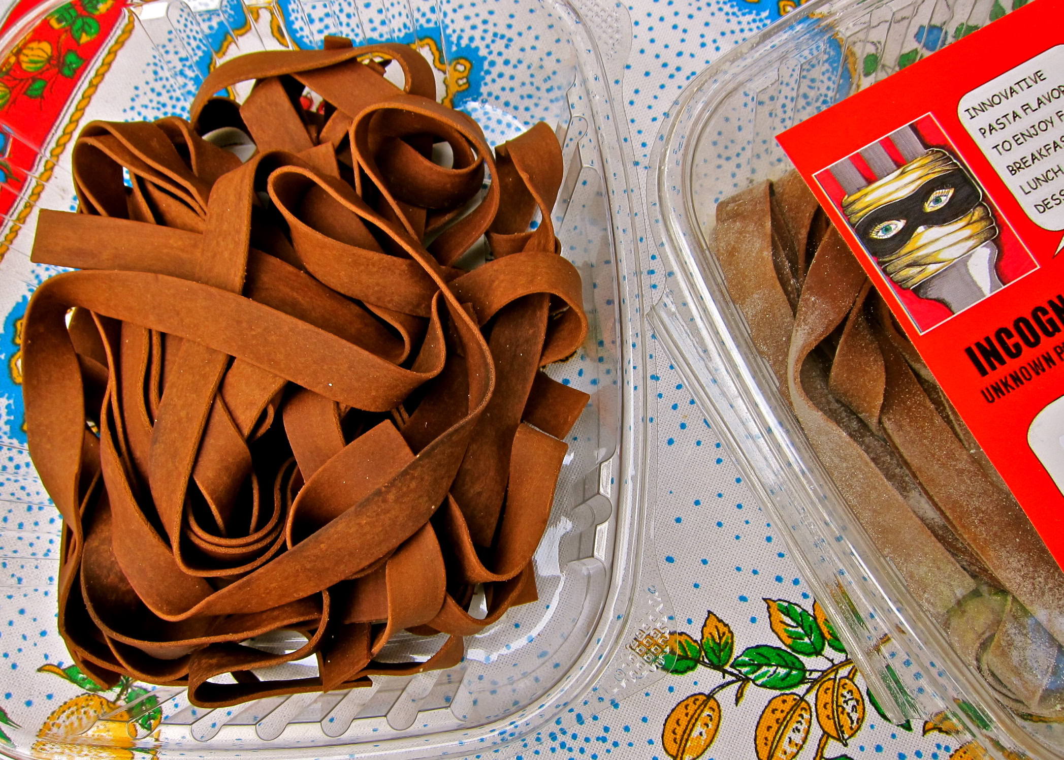 Close-up of brown pasta in a clear container with a red label, on a colorful tablecloth.