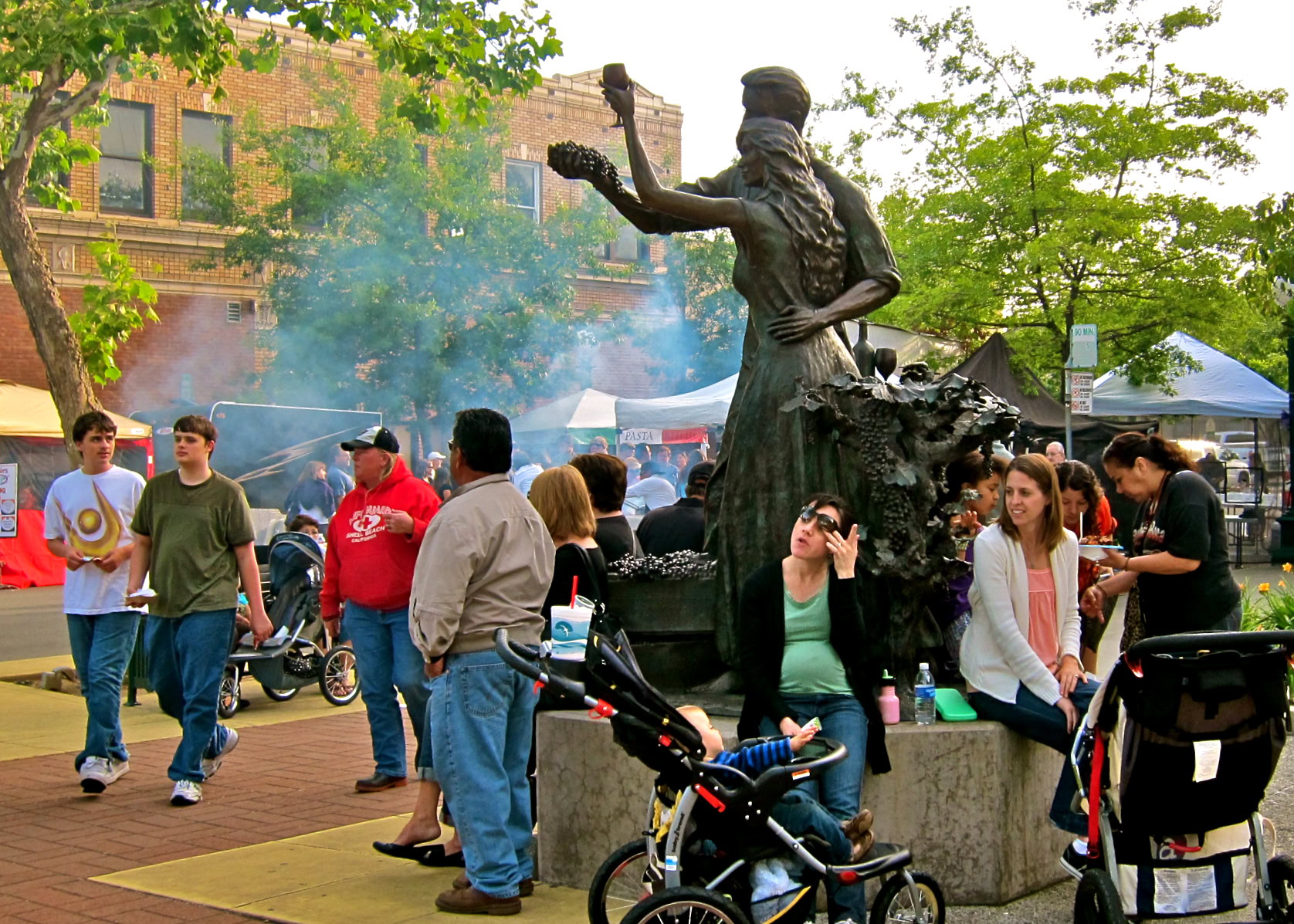 People gather around a statue at a lively outdoor event in Lodi, California, with trees and tents in the background.