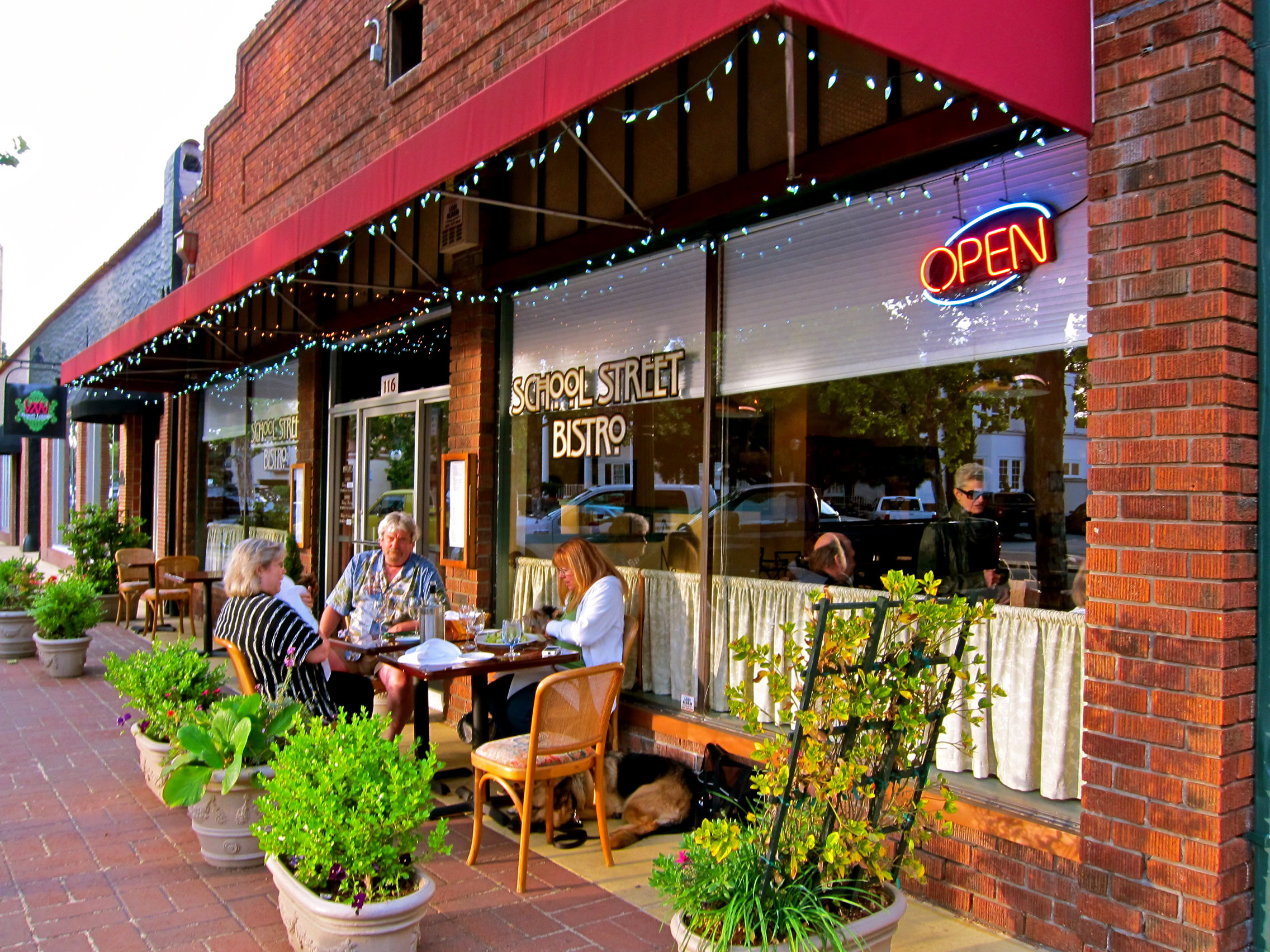 Outdoor dining scene at a Lodi winery with customers enjoying wine and food, surrounded by potted plants and festive stringed.