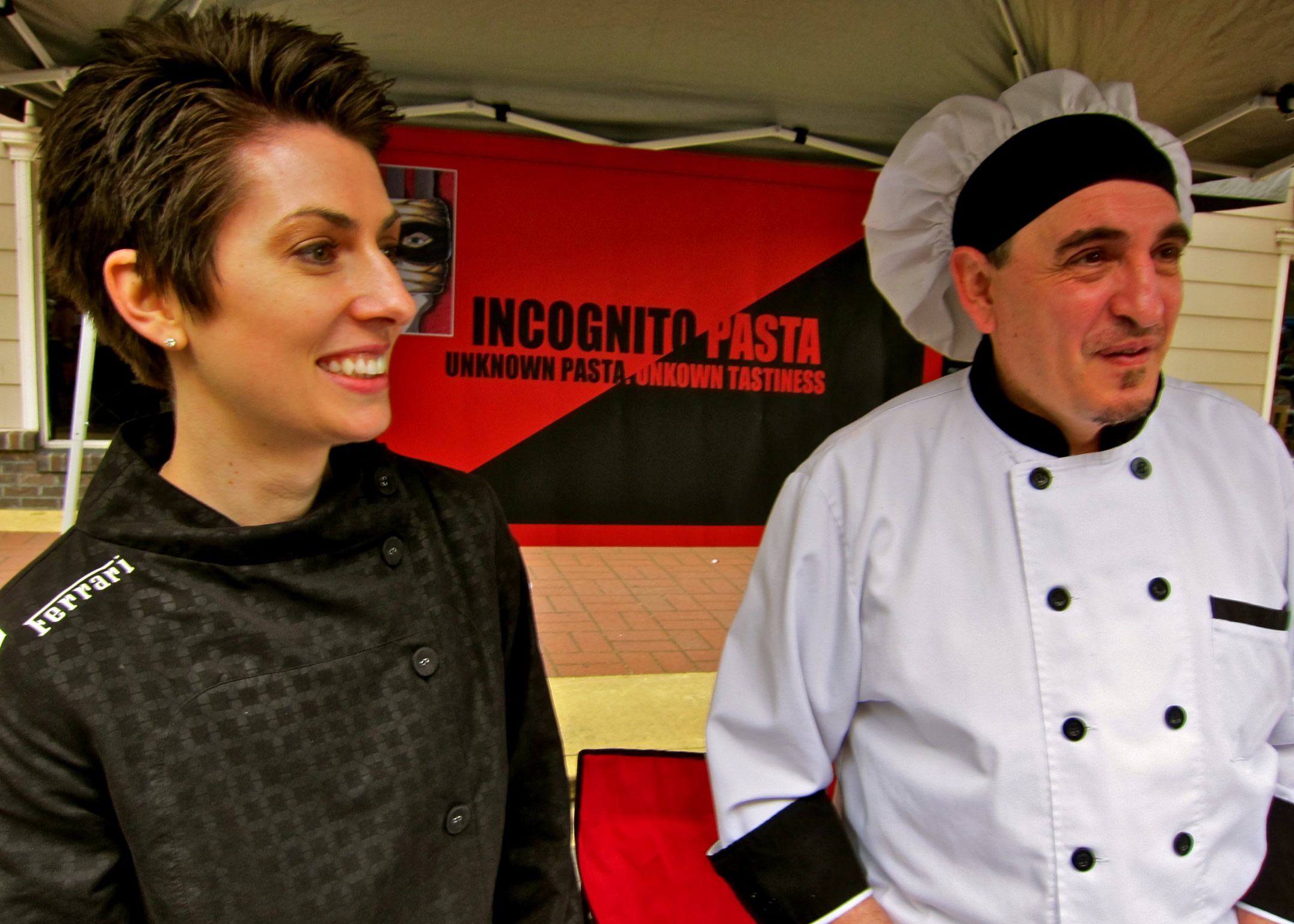 Two people at a wine event, one woman in a black chef's coat and one man in a white chef's uniform, smiling and talking.
