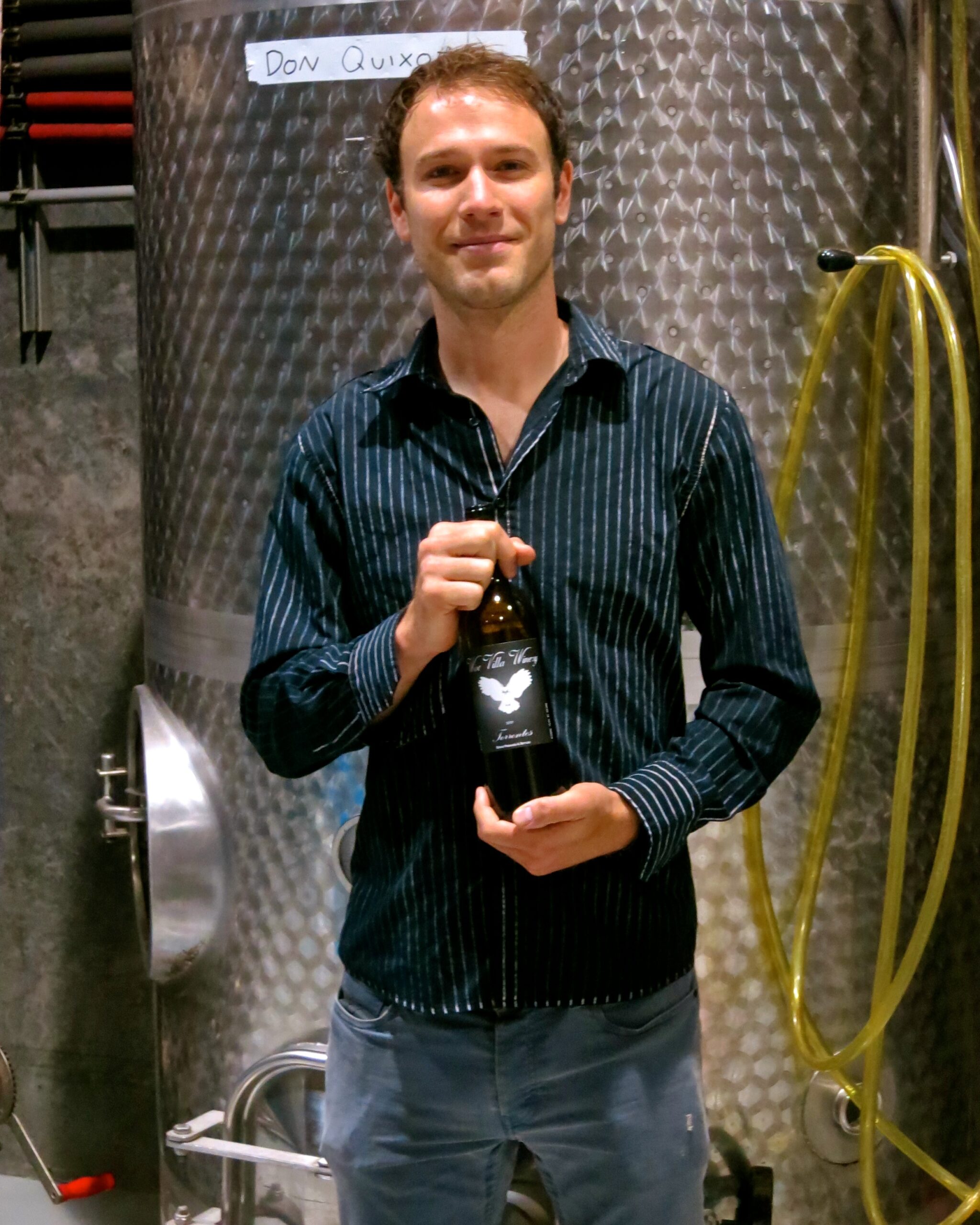 Man holding a wine bottle at a winery with stainless steel tanks in the background.