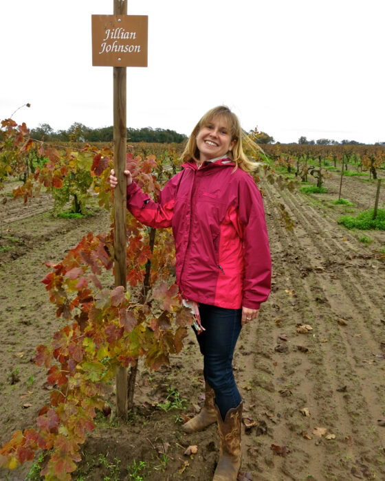 Smiling woman in pink jacket standing in vineyard with sign reading 'Jillian Johnson'.