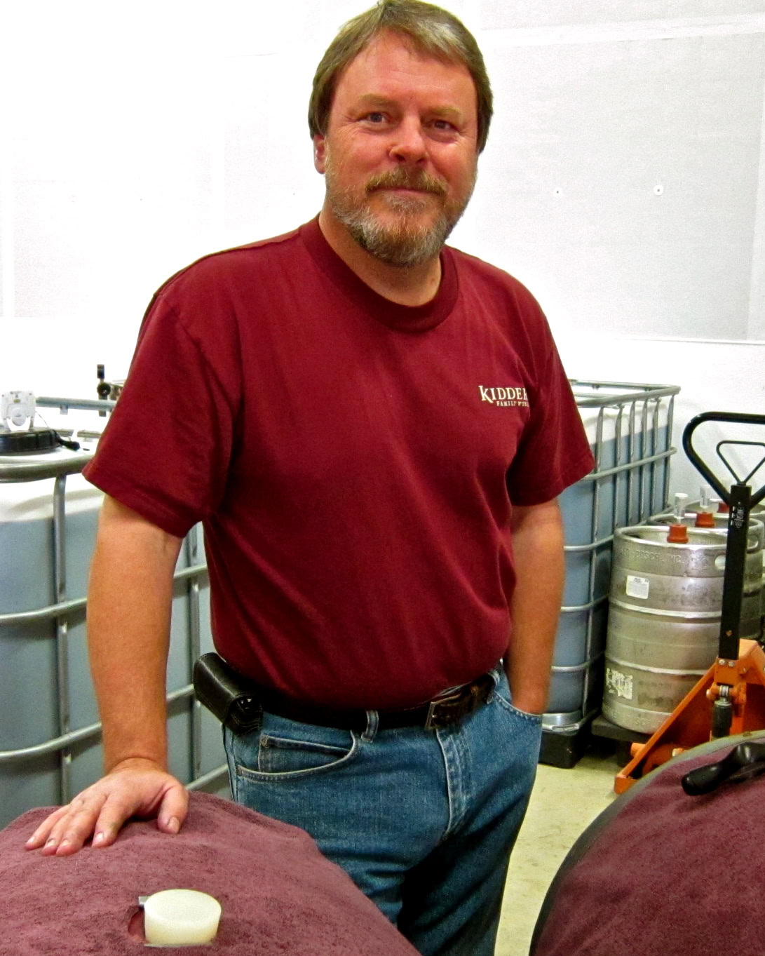 Man in red shirt at wine production facility with tanks and equipment in background.