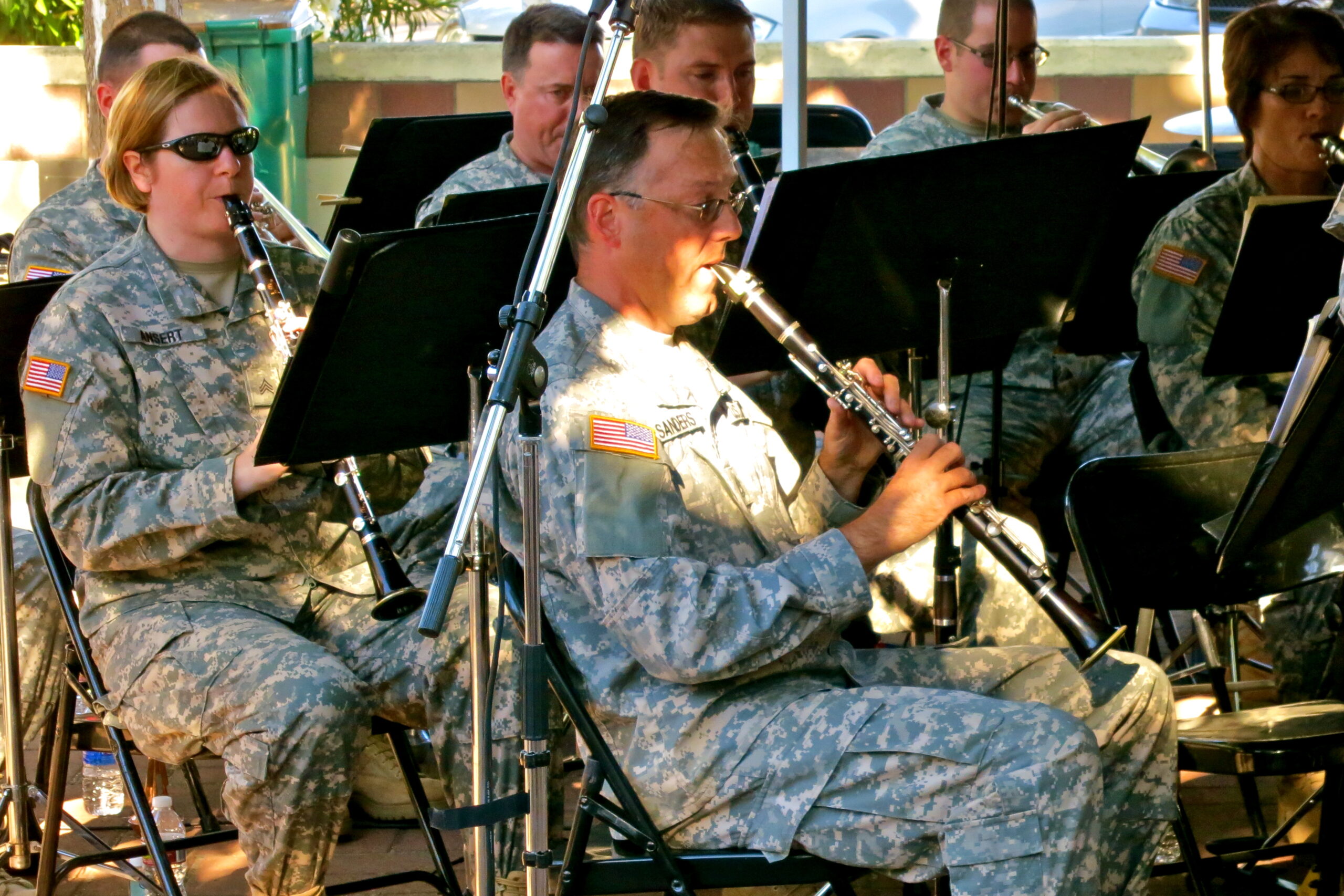 Military band performing outdoors with musicians in camouflage uniforms and musical instruments.