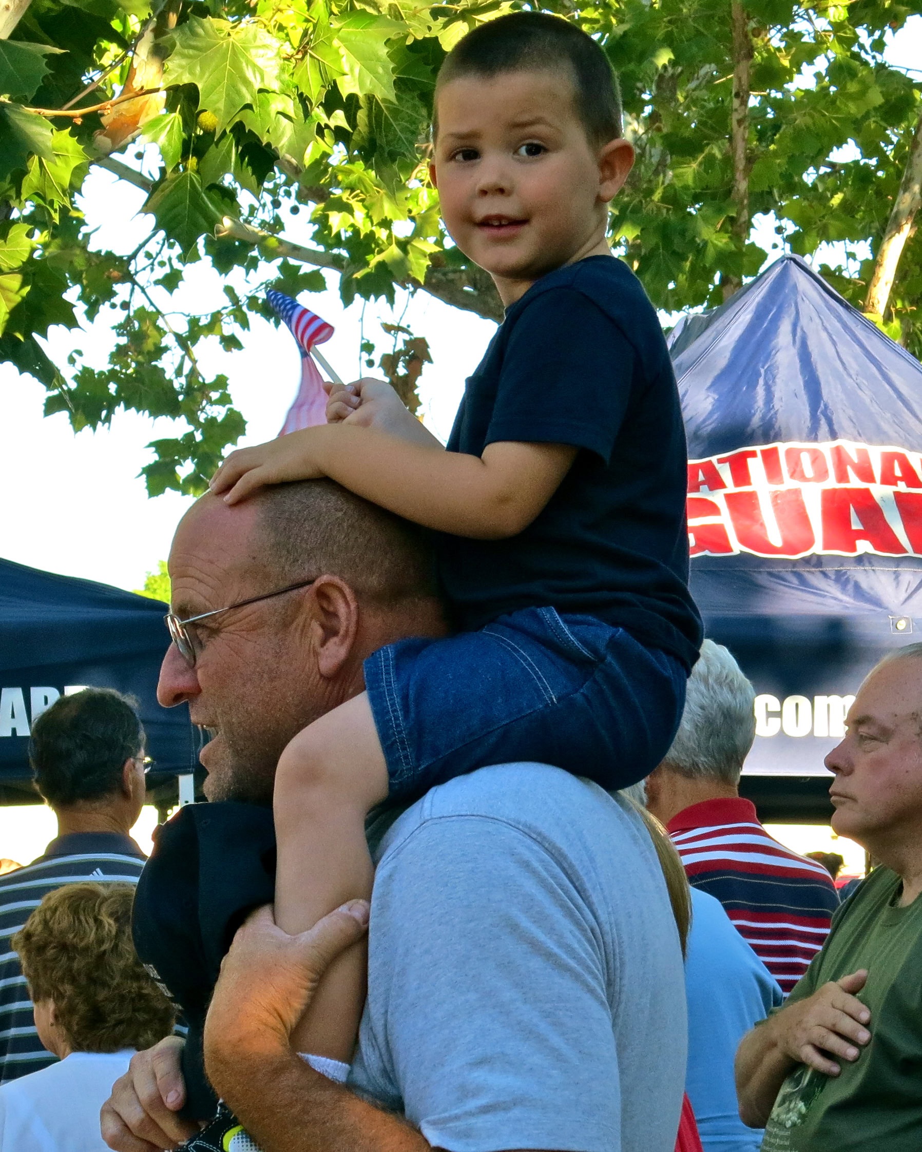 Child sitting on man's shoulders at Lodi wine event with crowd and trees in background.