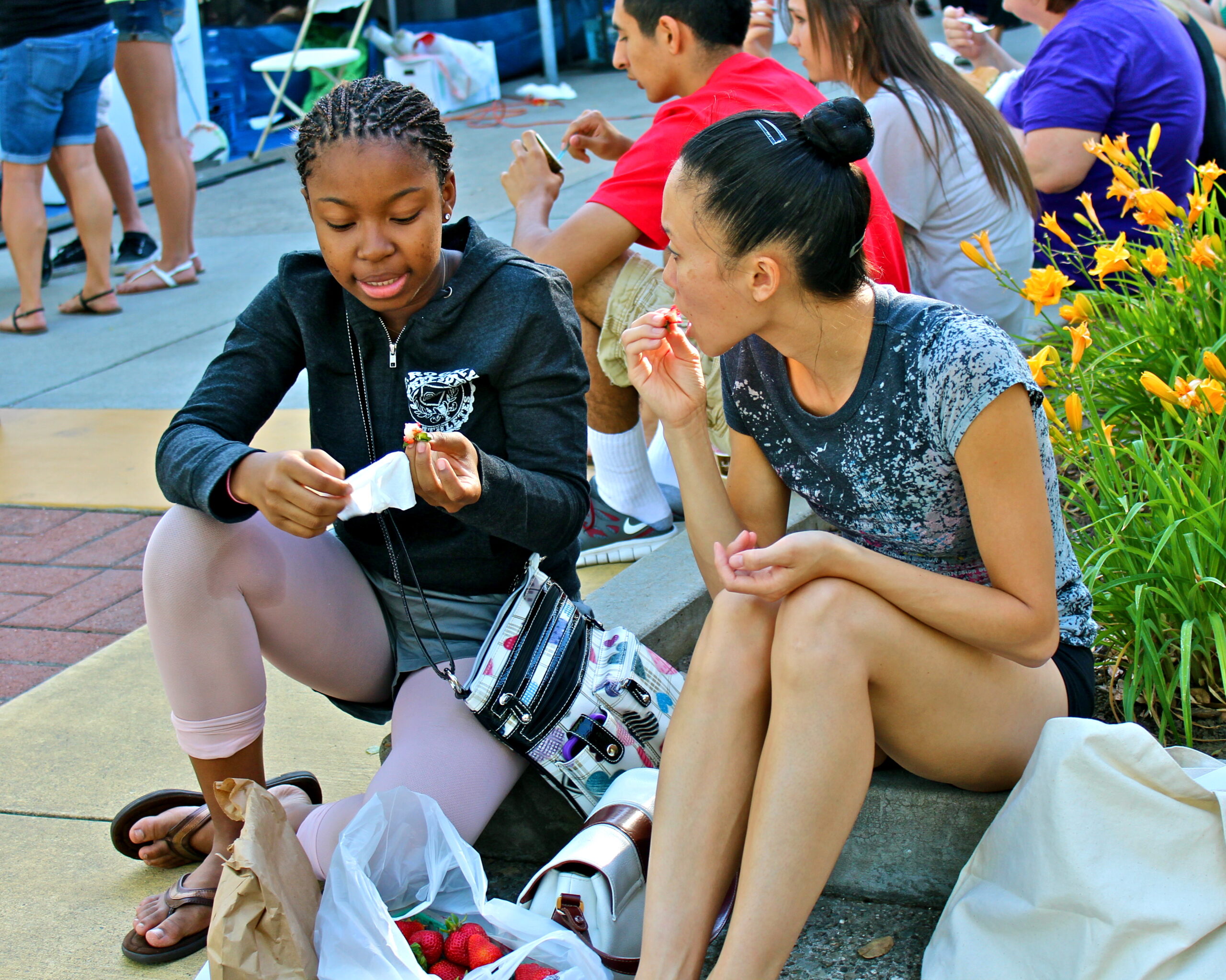 Two women sitting outdoors, enjoying a snack at a Lodi wine event, with other attendees in the background.