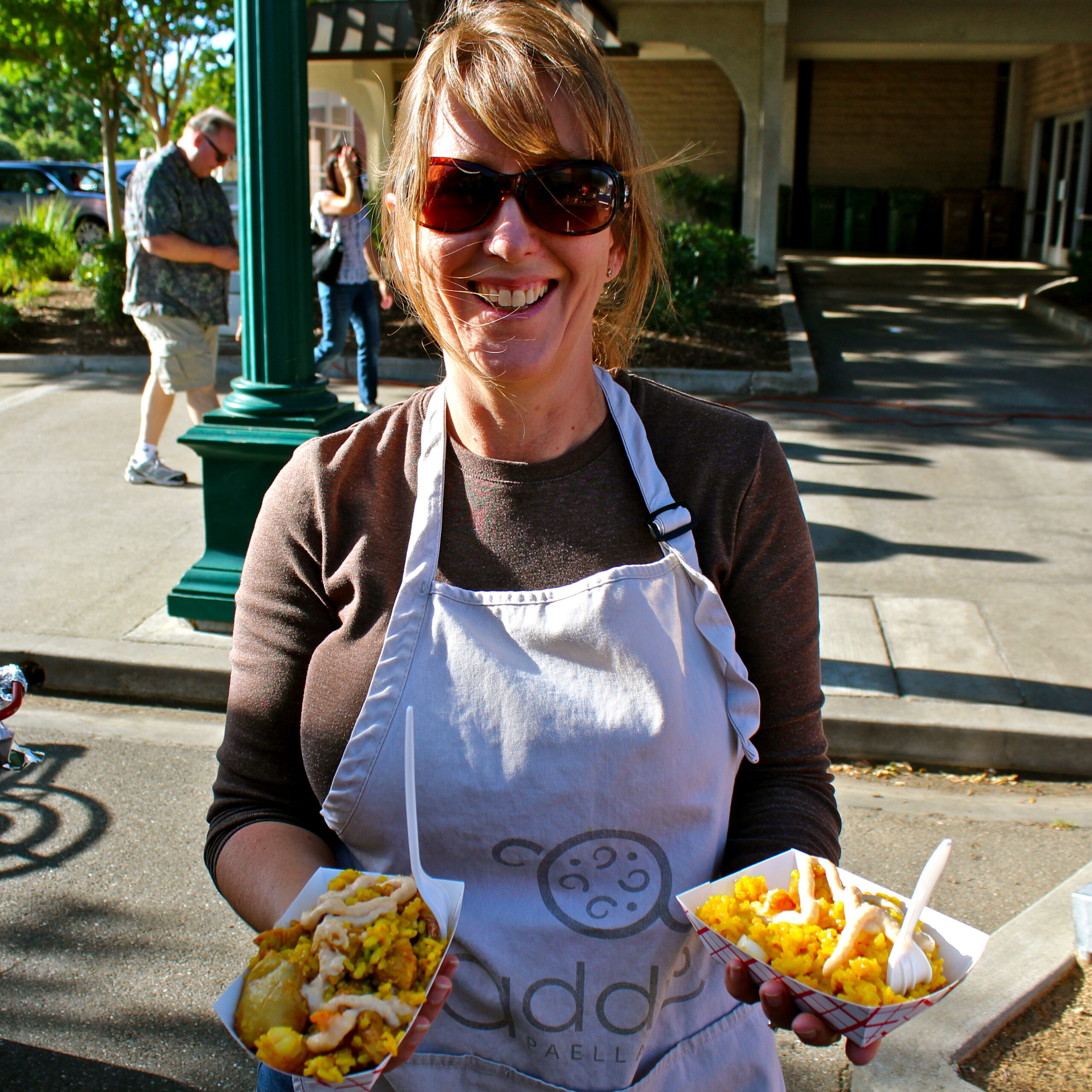 Smiling woman in apron holding food at Lodi Winegrape Commission event outdoors.