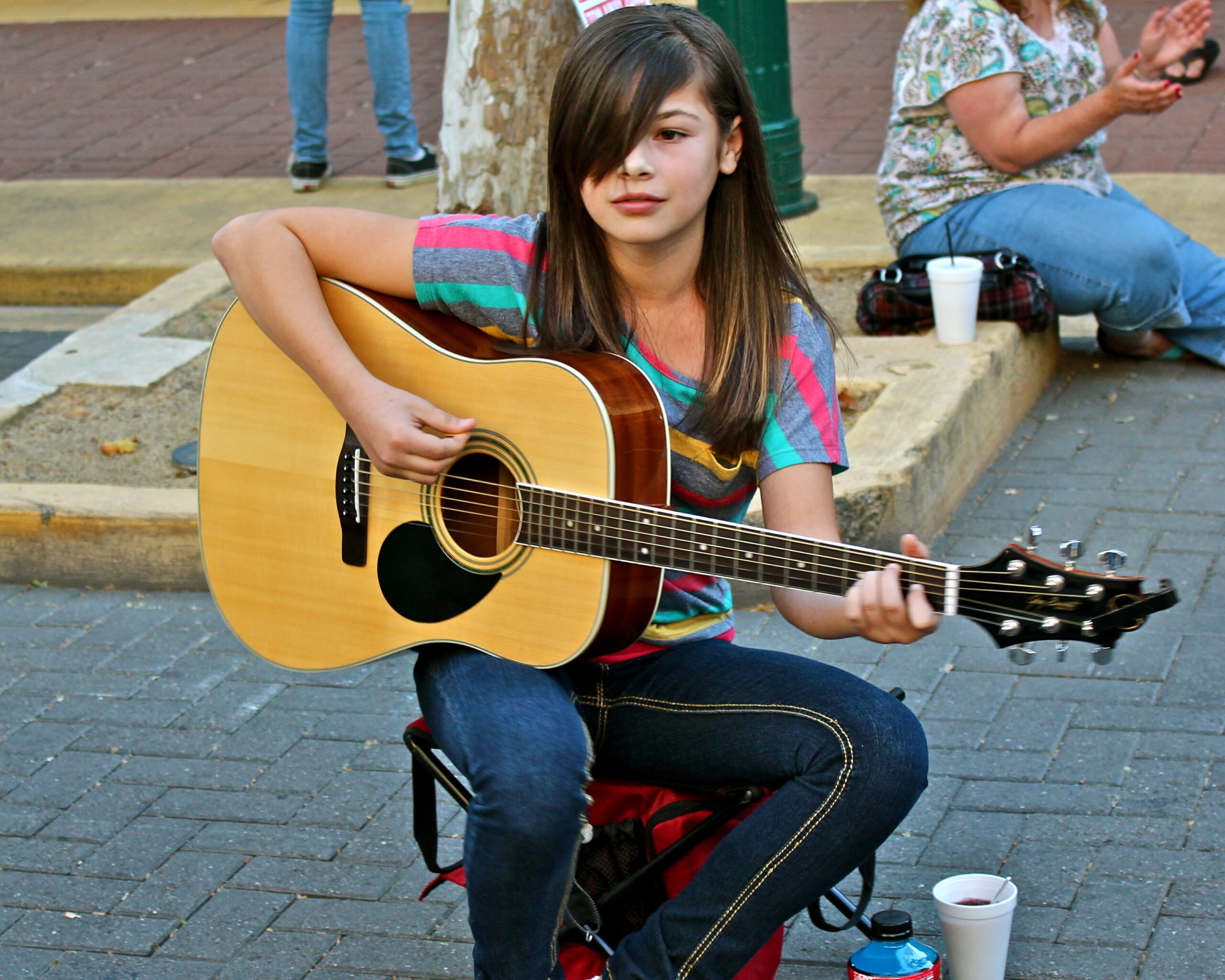 Young girl playing acoustic guitar outdoors in Lodi, California, with people sitting nearby.