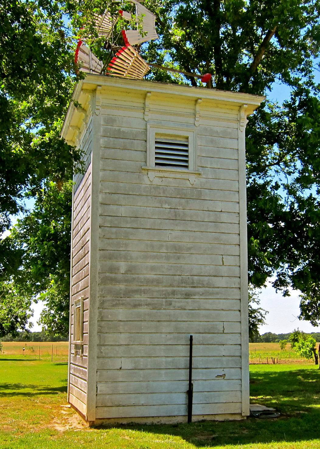 Historic wooden tower with weather vane and antennas in Lodi, California, surrounded by green trees and open fields.