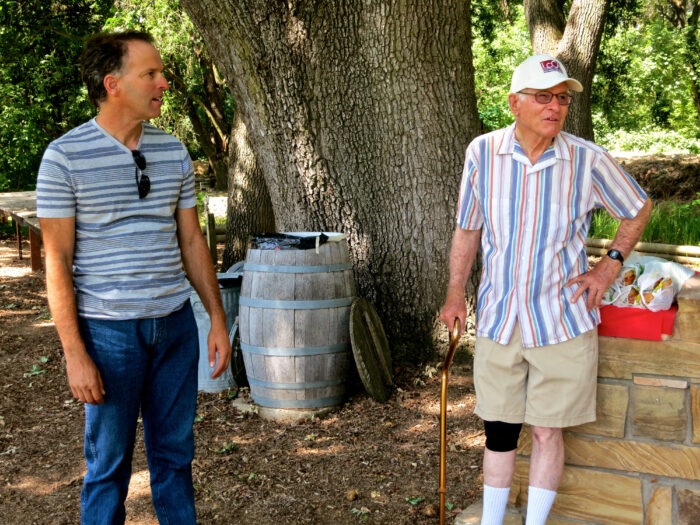 Two men talking outdoors near a large tree, wine barrel, and picnic area, representing Lodi Winegrape Commission activities.