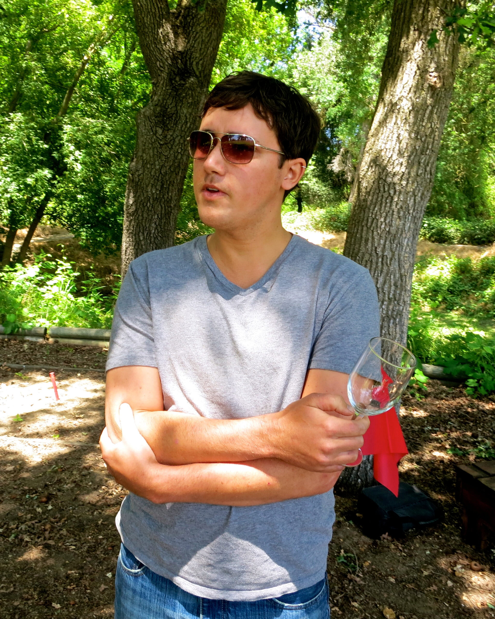 Man with sunglasses holding a wine glass outdoors in a lush green setting.