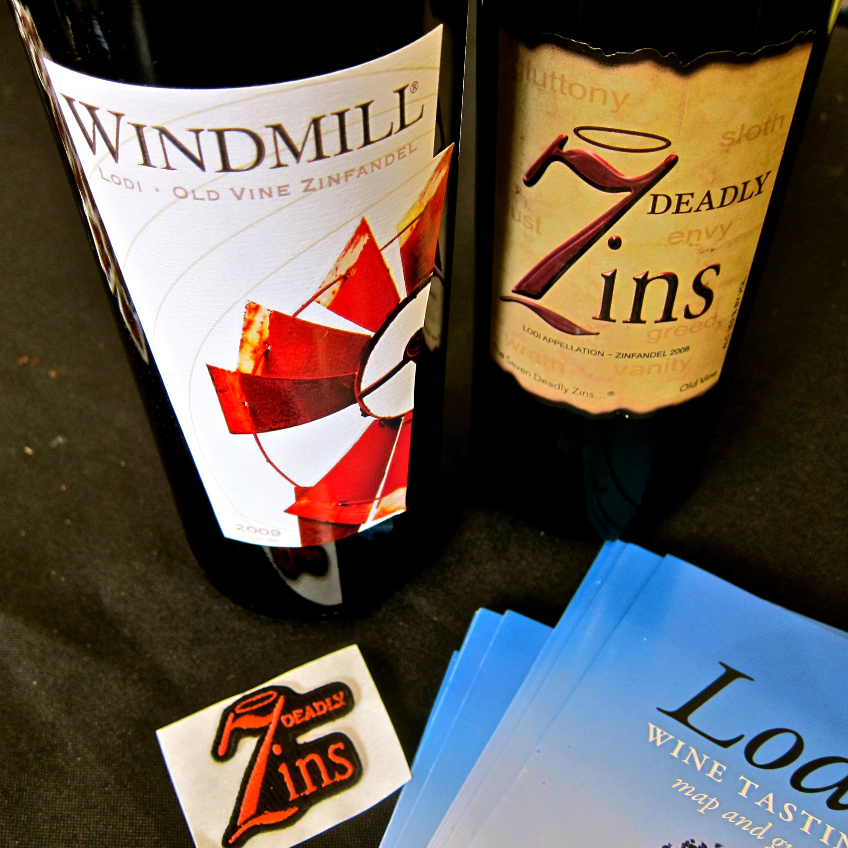 Two wine bottles from Lodi Winegrape Commission, Windmill and Deadly Zins, with promotional materials on a dark surface.