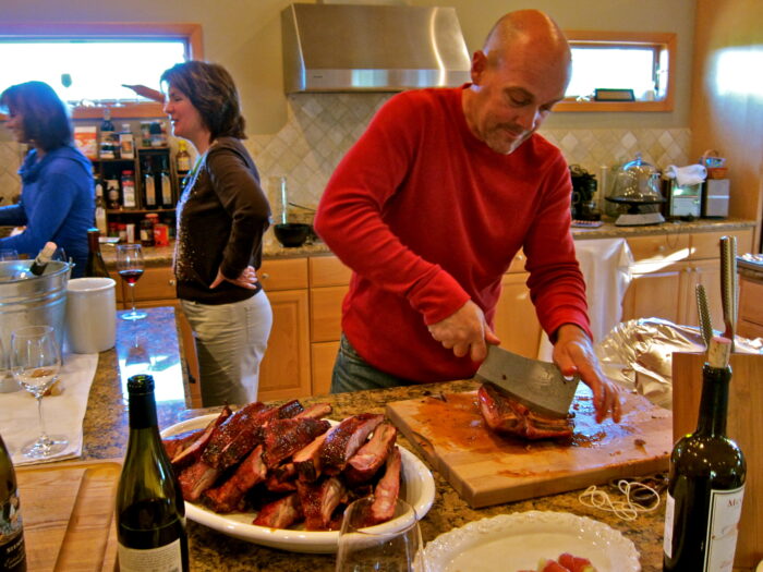 Man slicing cooked meat at a social gathering with wine bottles and people in the background.