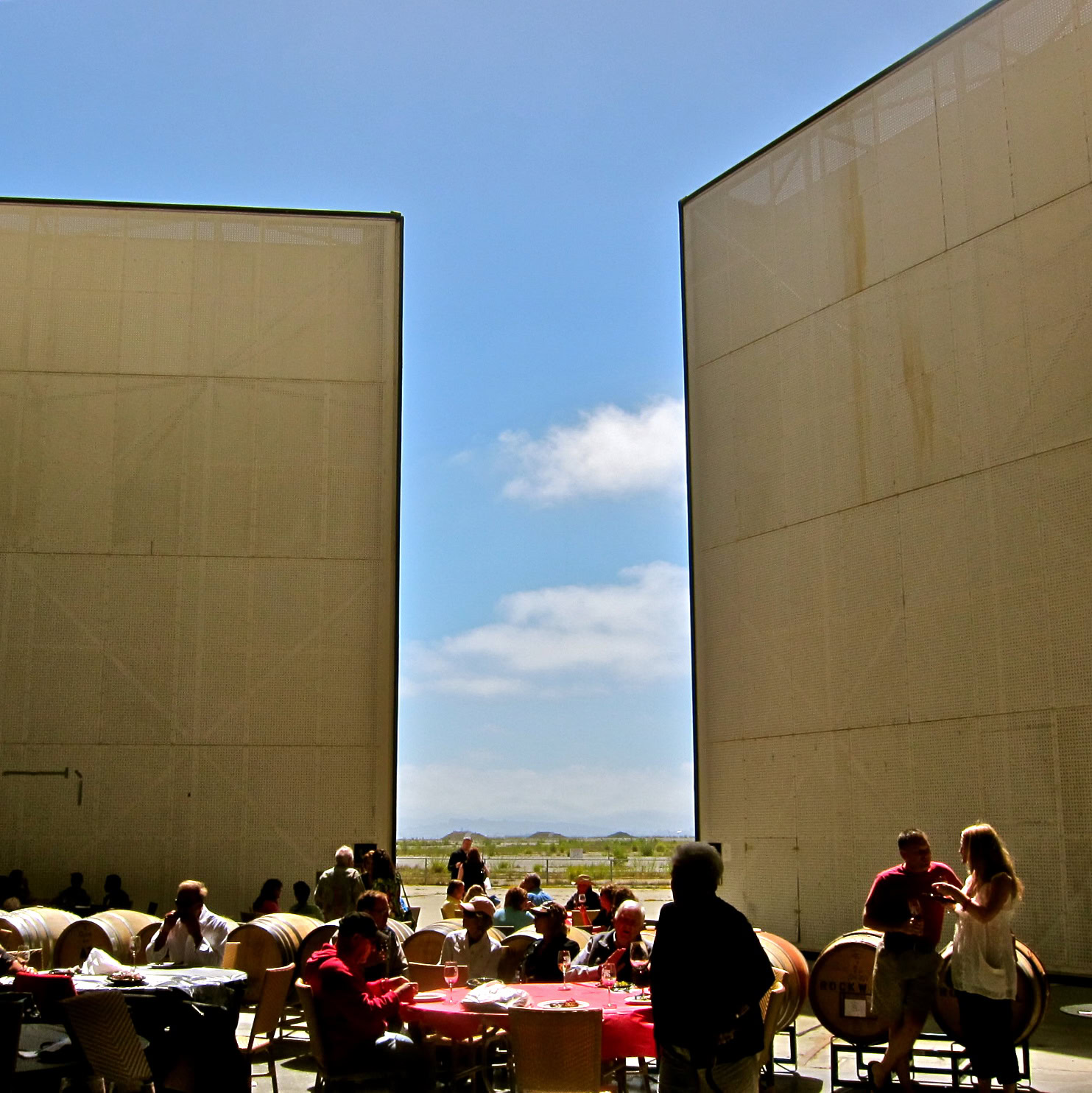 People enjoying outdoor wine tasting event between large beige walls under a blue sky.
