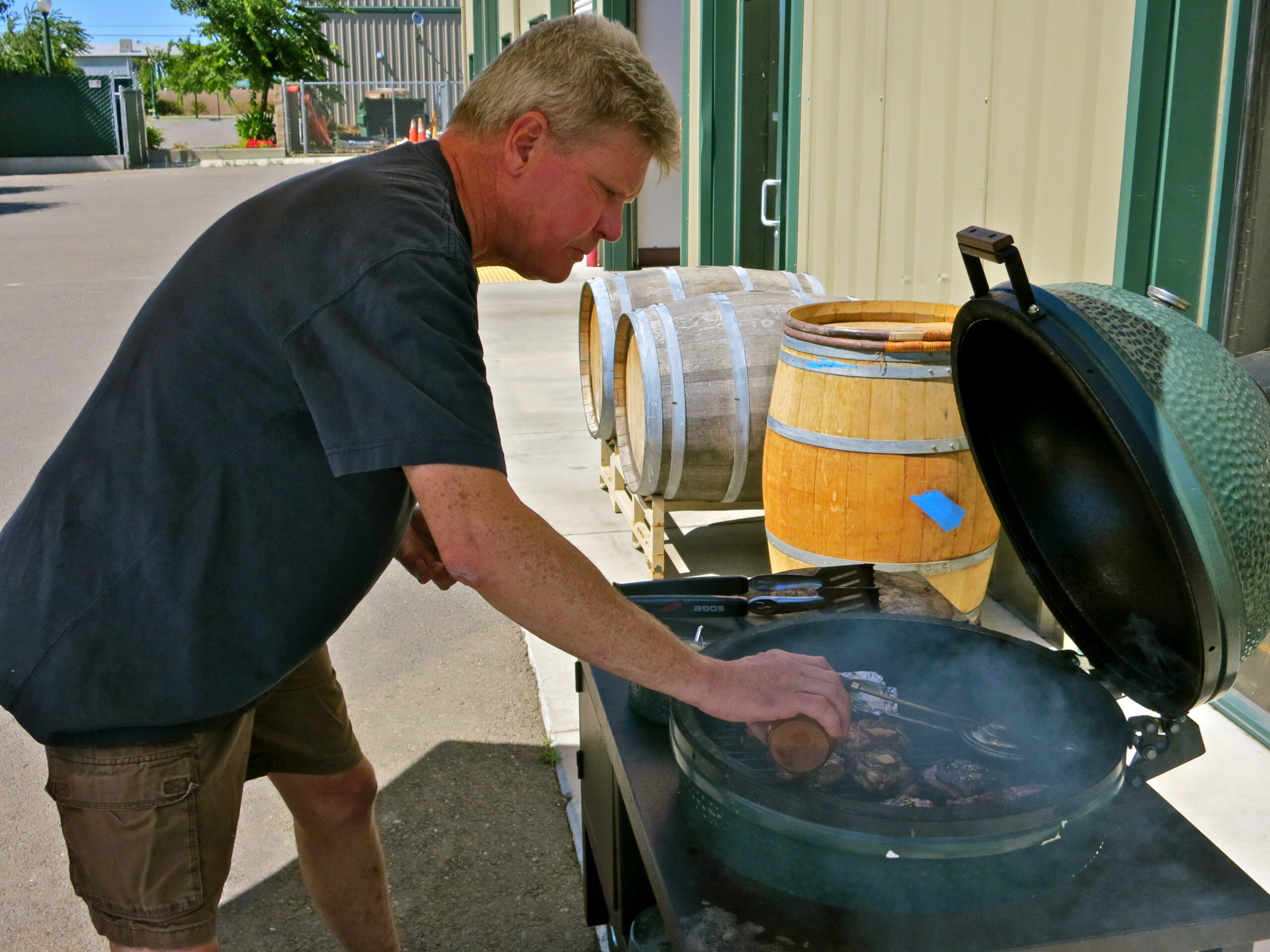 Man grilling steaks outdoors near wine barrels at Lodi Winegrape Commission event.