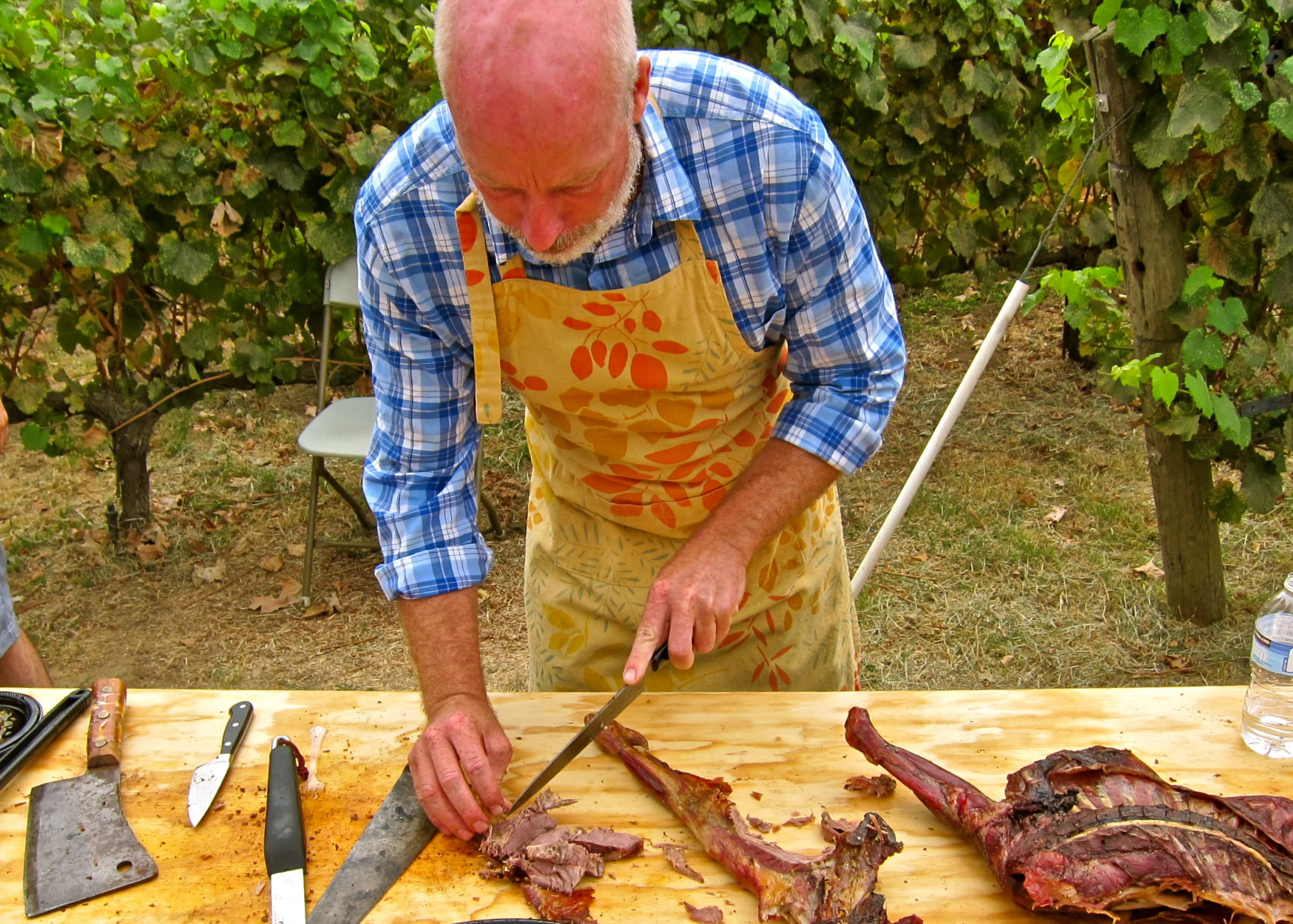 Man preparing and slicing cooked meat outdoors in a vineyard setting for wine pairing.