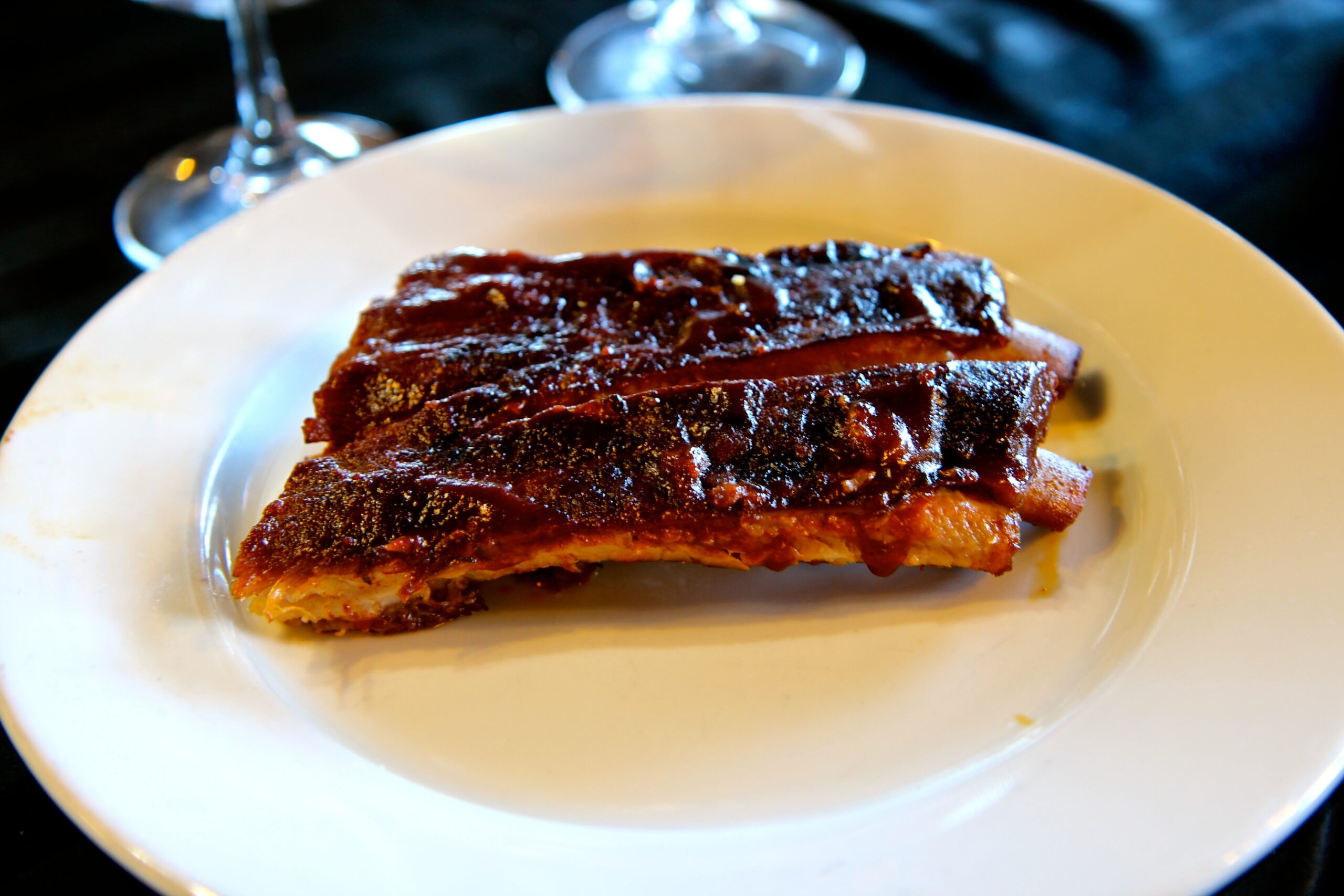 Close-up of a glazed pork rib on a white plate, with wine glasses in the background.