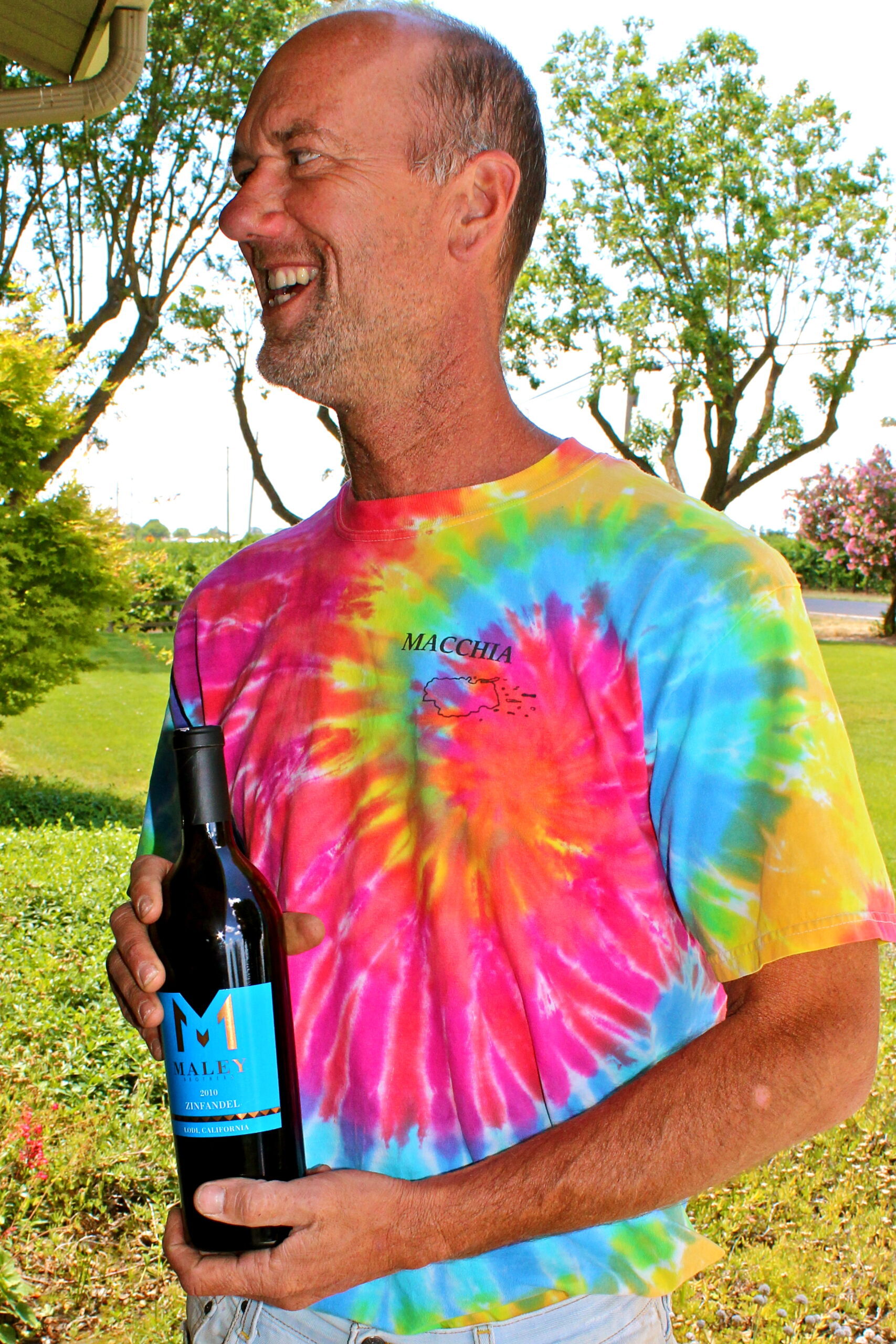 Smiling man in tie-dye shirt holding a wine bottle outdoors at a Lodi wine event.