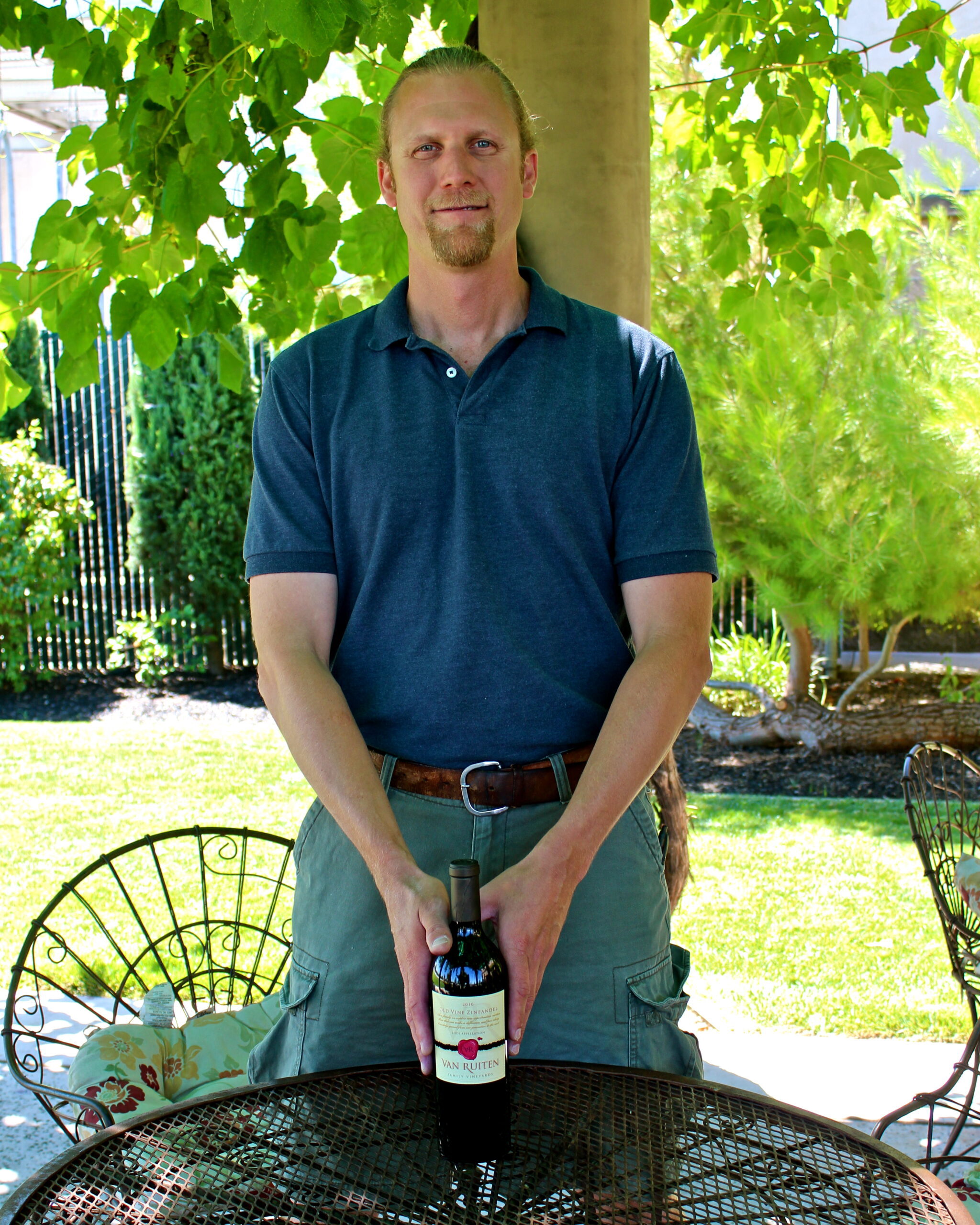 Man holding a bottle of wine outdoors in a vineyard setting with lush green foliage.