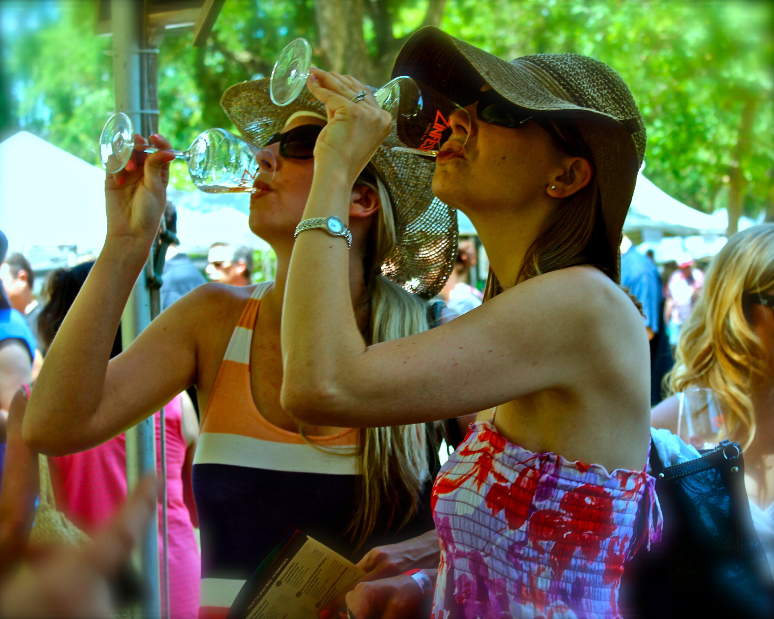 Two women enjoying wine at an outdoor festival, wearing hats and sunglasses, with trees and tents in the background.