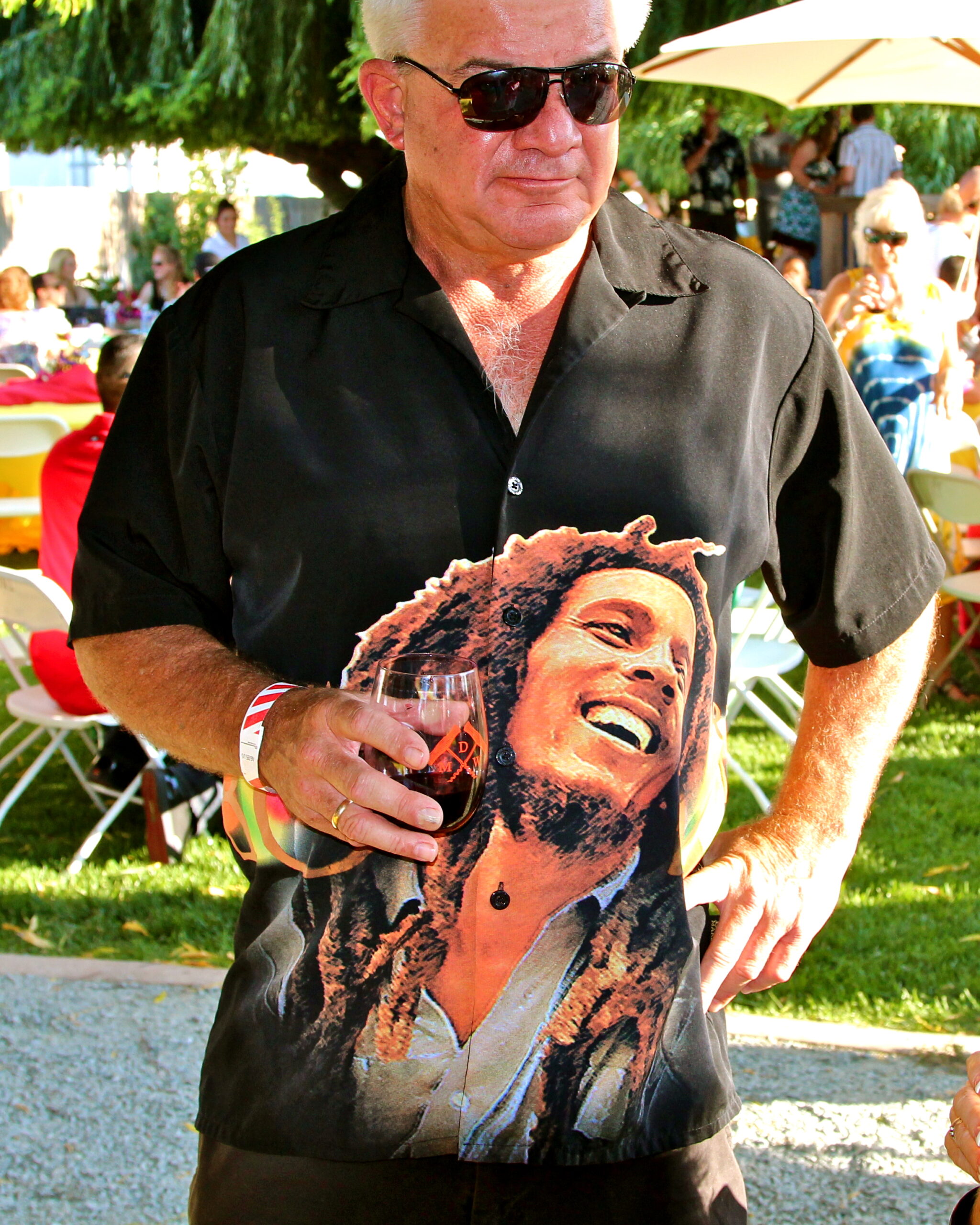 Man wearing a black shirt with Bob Marley image holding a glass of red wine at outdoor event.