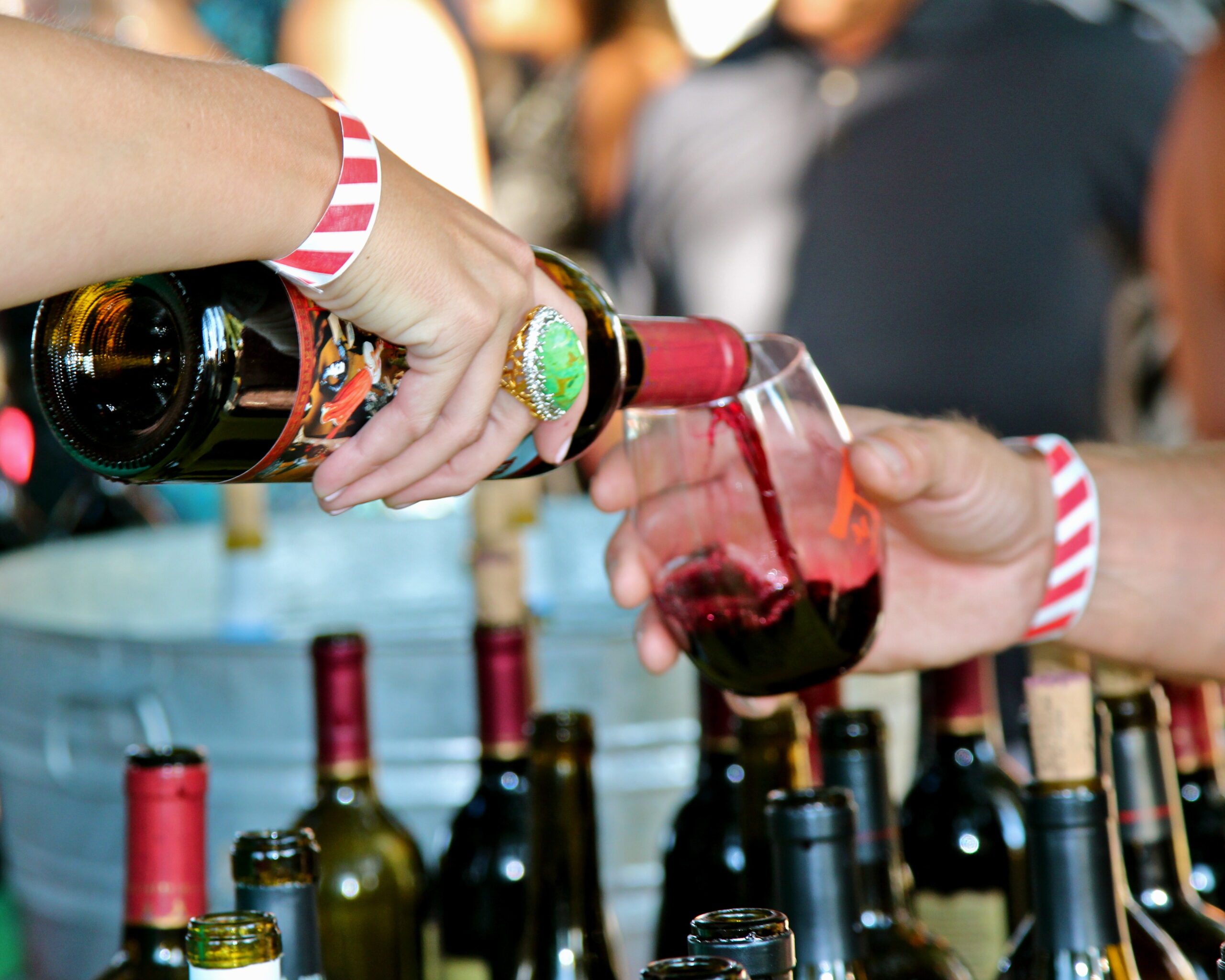 Person pouring red wine into glass at a wine tasting event, with wine bottles on table.