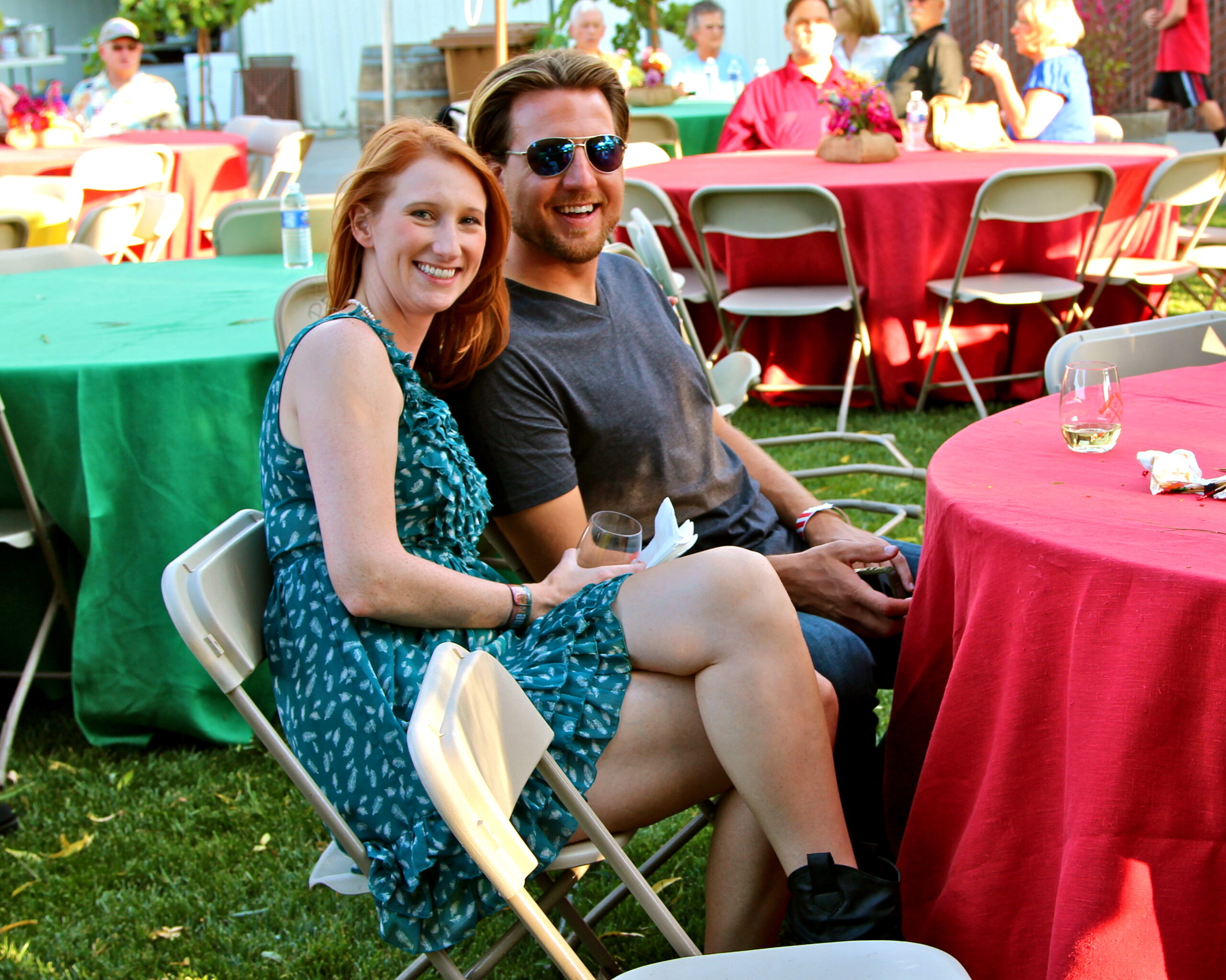 Smiling couple enjoying wine at an outdoor event with colorful tables and guests in the background.