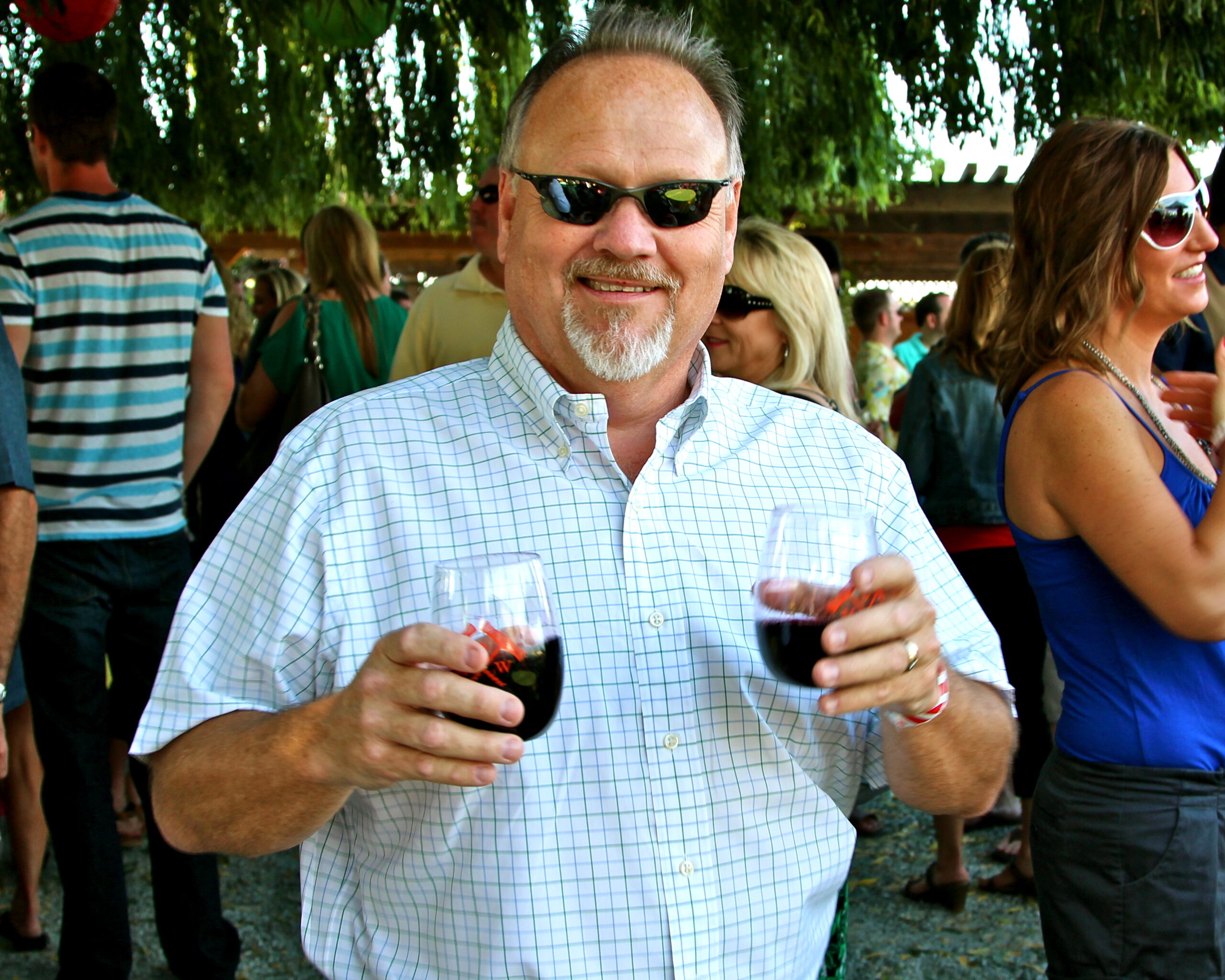 Smiling man with sunglasses holding two glasses of red wine at a wine tasting event.