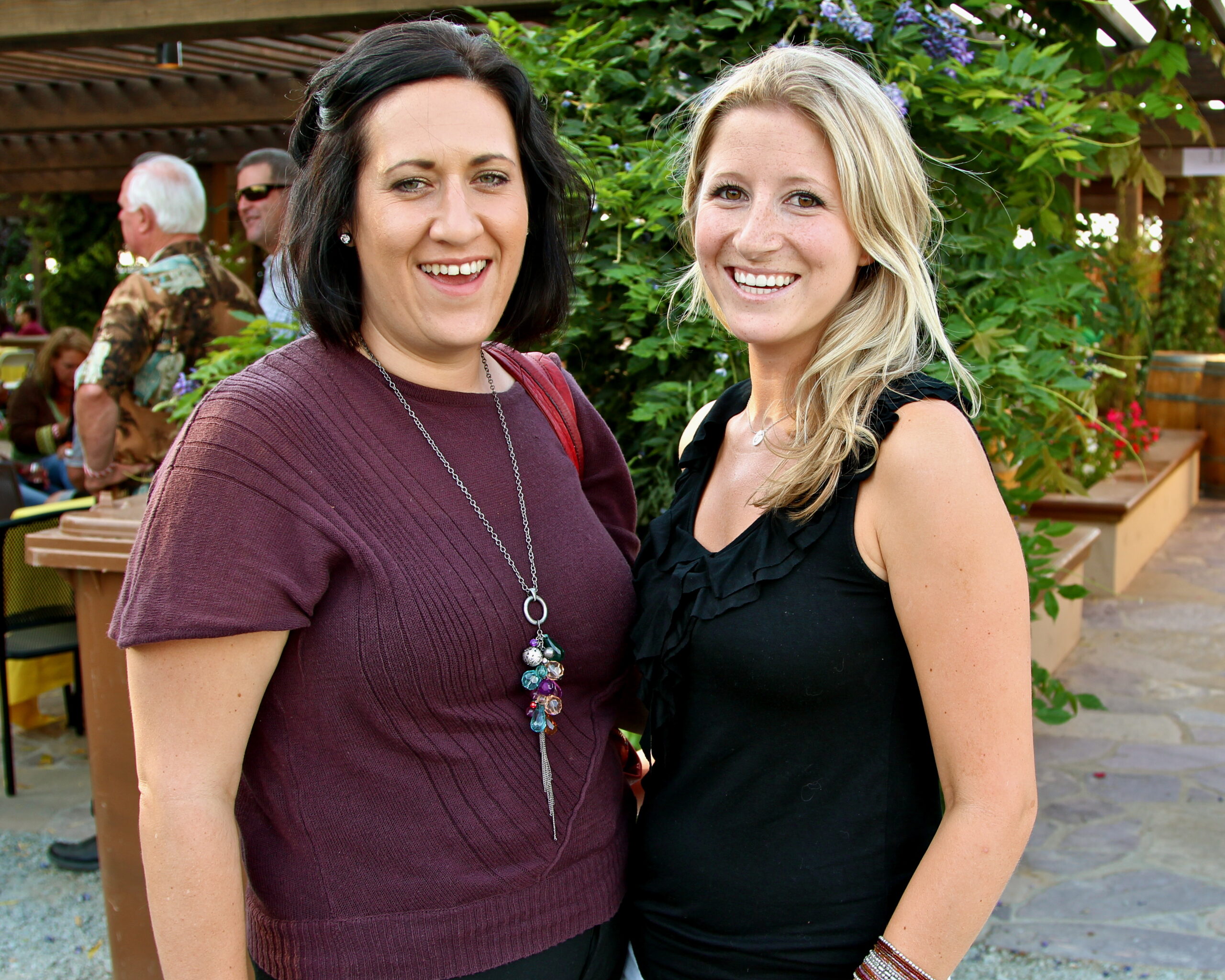 Two women smiling at a Lodi Winegrape Commission event outdoors with greenery in the background.