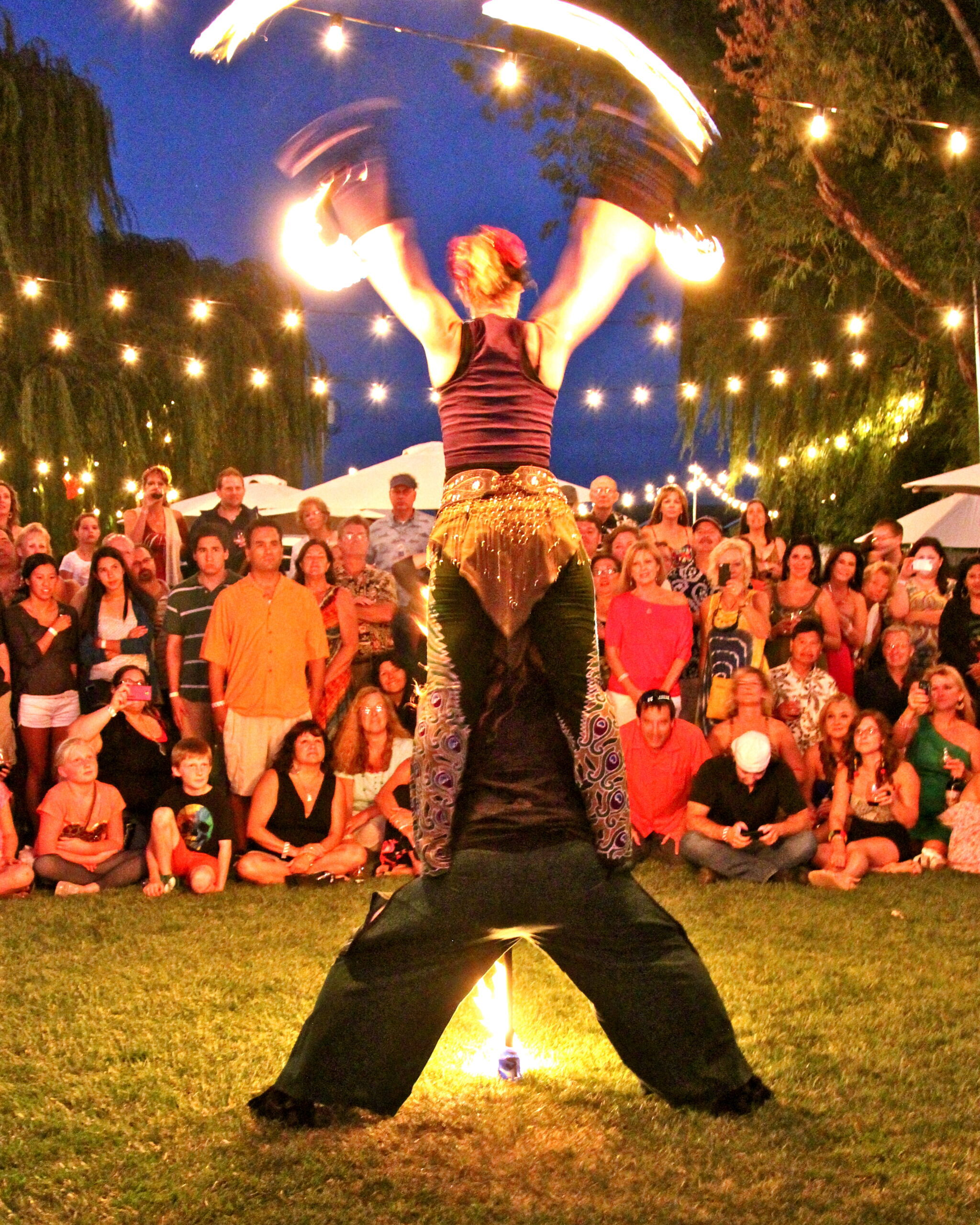 Performer spinning fire at outdoor event with audience and string lights.