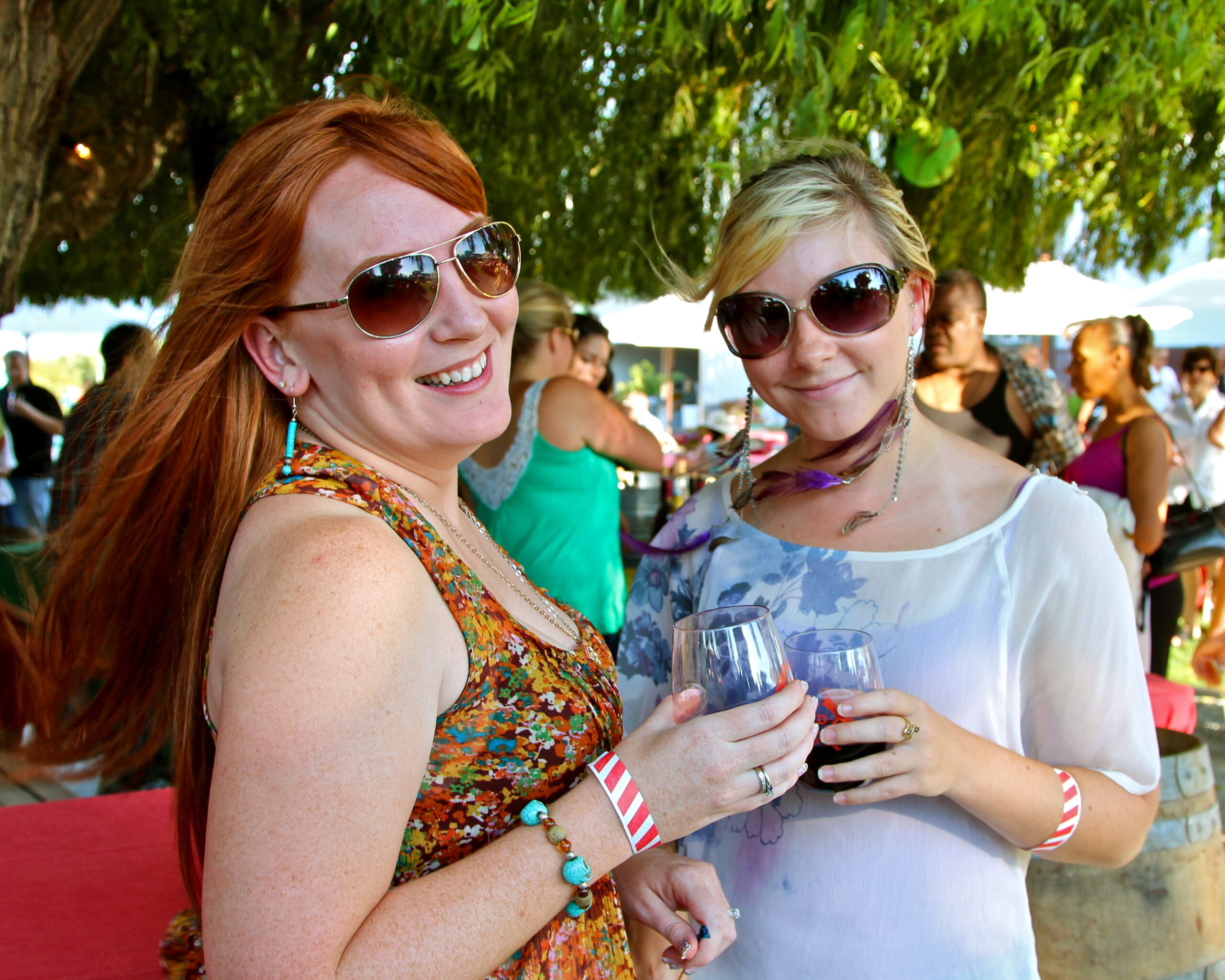 Two women enjoying wine at a Lodi Winegrape Commission event outdoors with trees and other guests in the background.