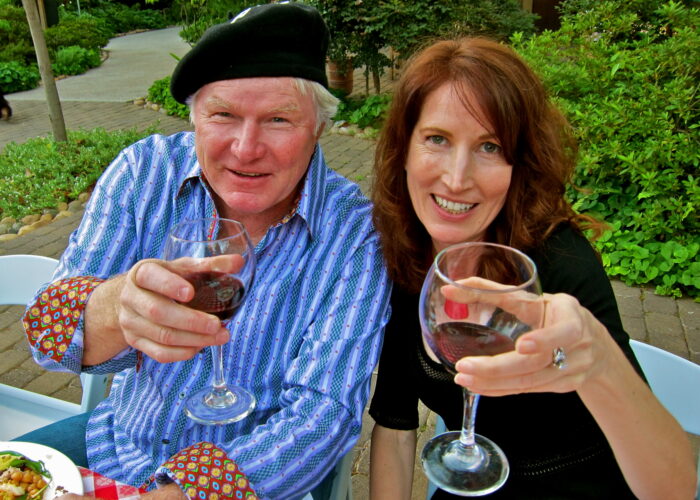 Two people enjoying wine outdoors, smiling and raising glasses of red wine, with lush greenery in the background.