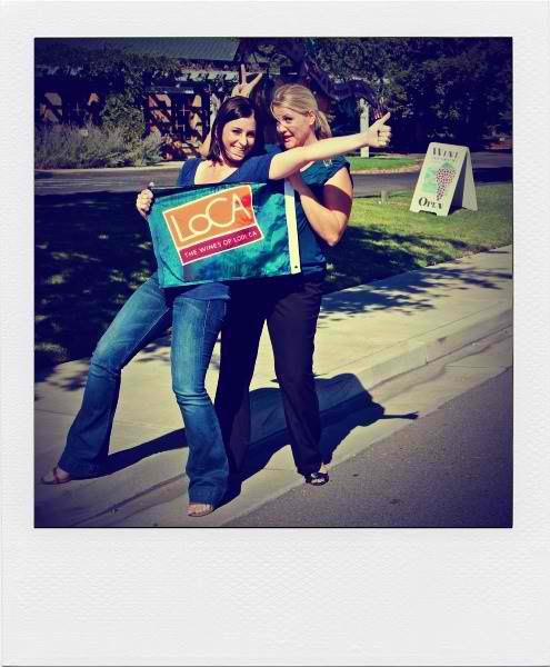 Two women holding a Lodi Winegrape Commission sign outdoors, smiling and taking a selfie.