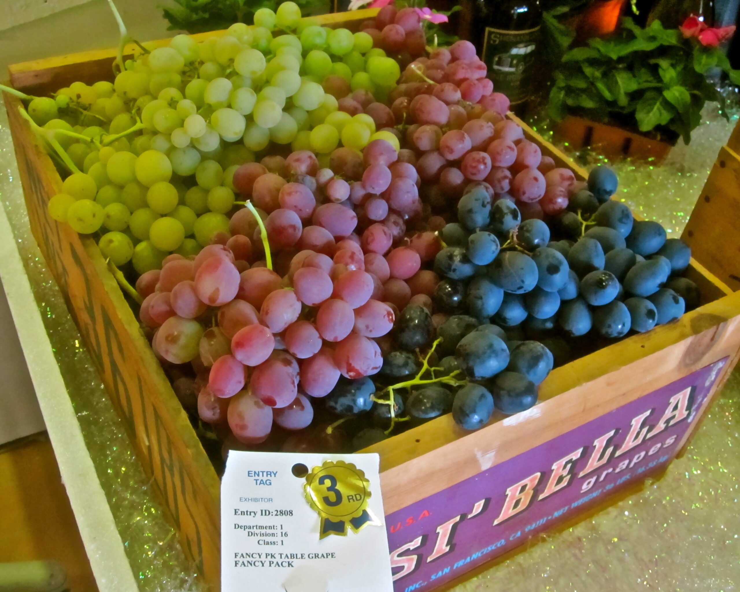 Colorful bunches of green, pink, and black grapes in a wooden crate at a market.