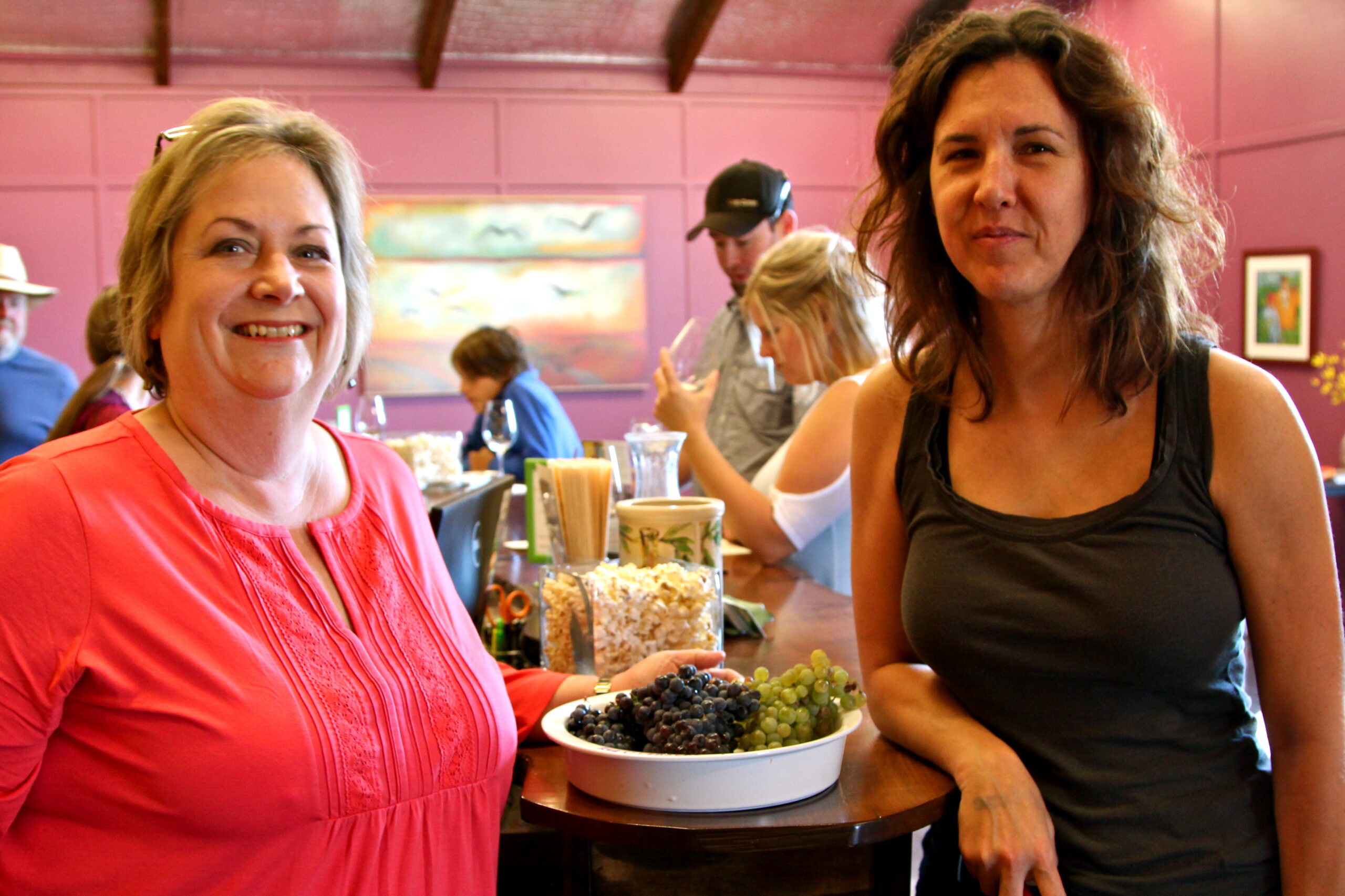 Two women at a wine tasting event with grapes and wine glasses in the background.