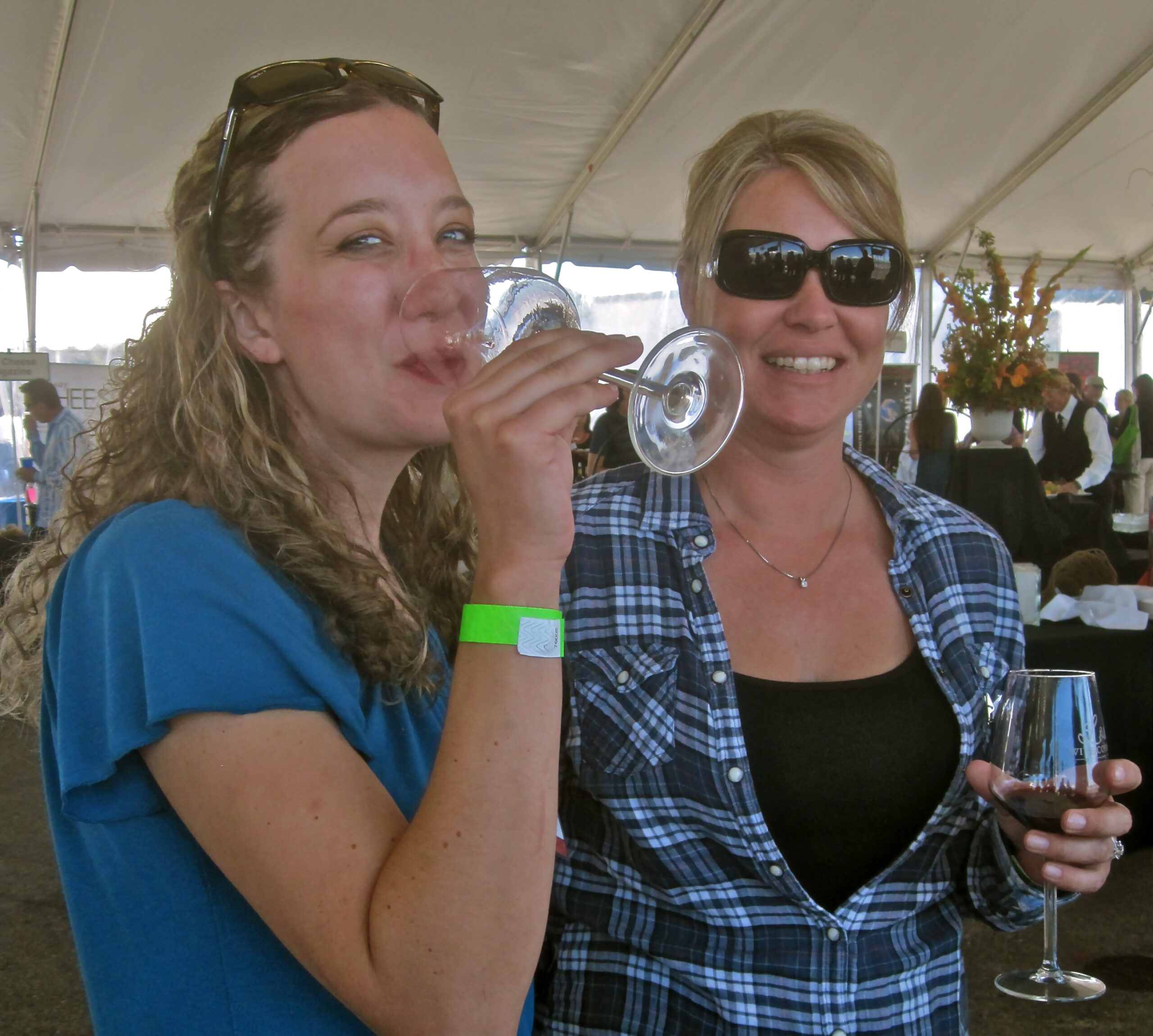 Two women enjoying wine at a wine tasting event under a large tent.