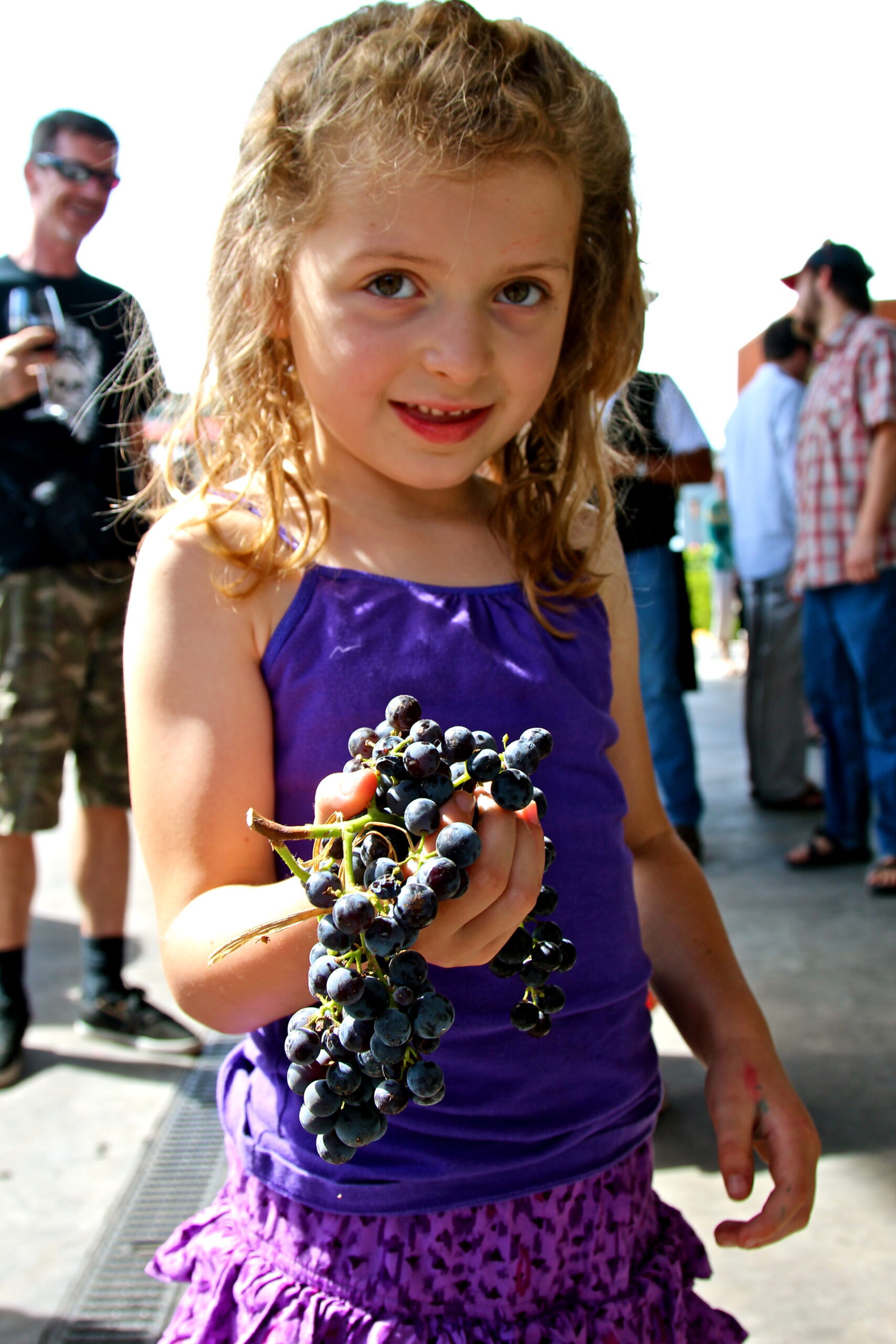 Young girl holding a bunch of dark grapes at a Lodi wine event with people in the background.