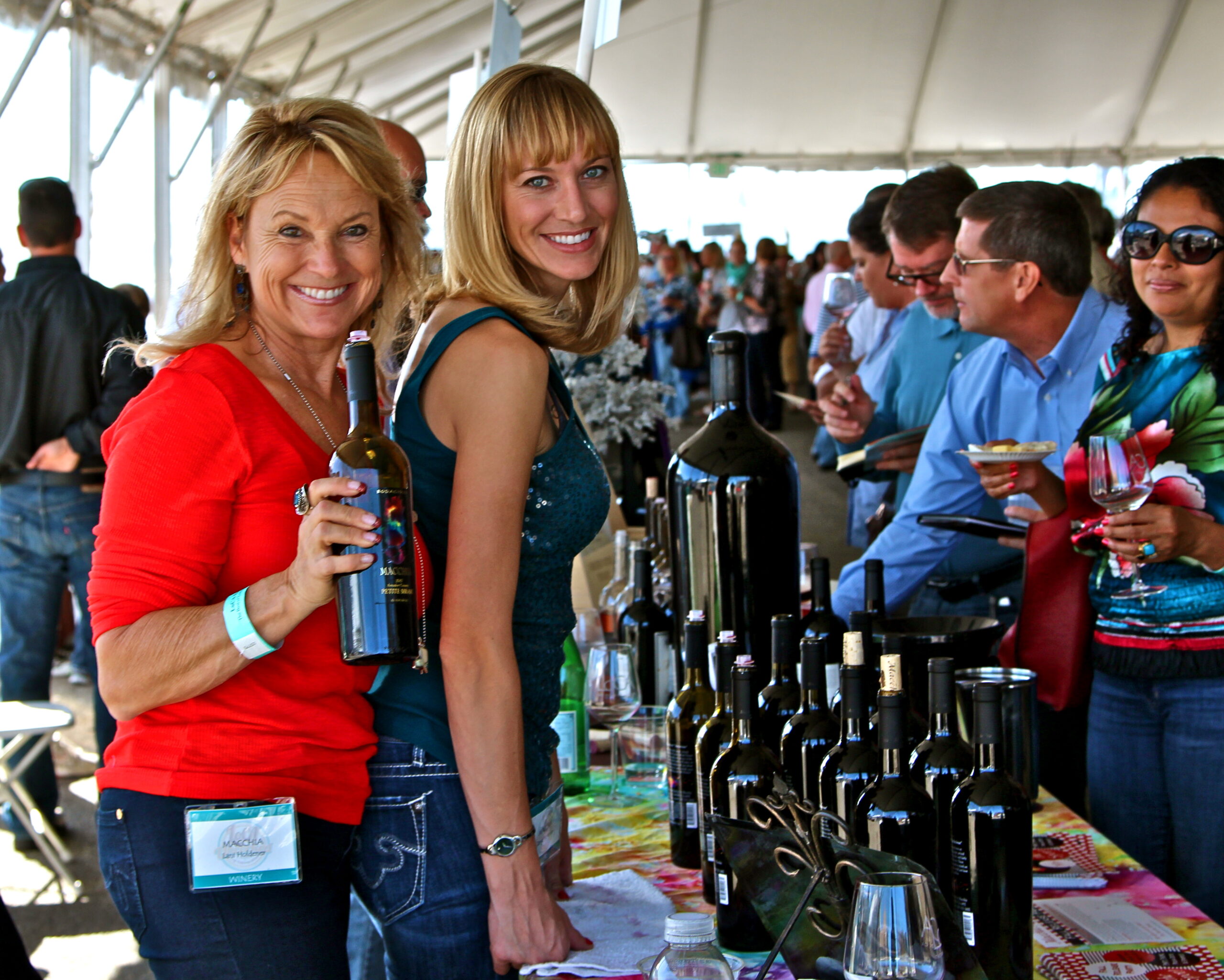 Smiling women at a wine tasting event with bottles of wine and a crowd in the background.