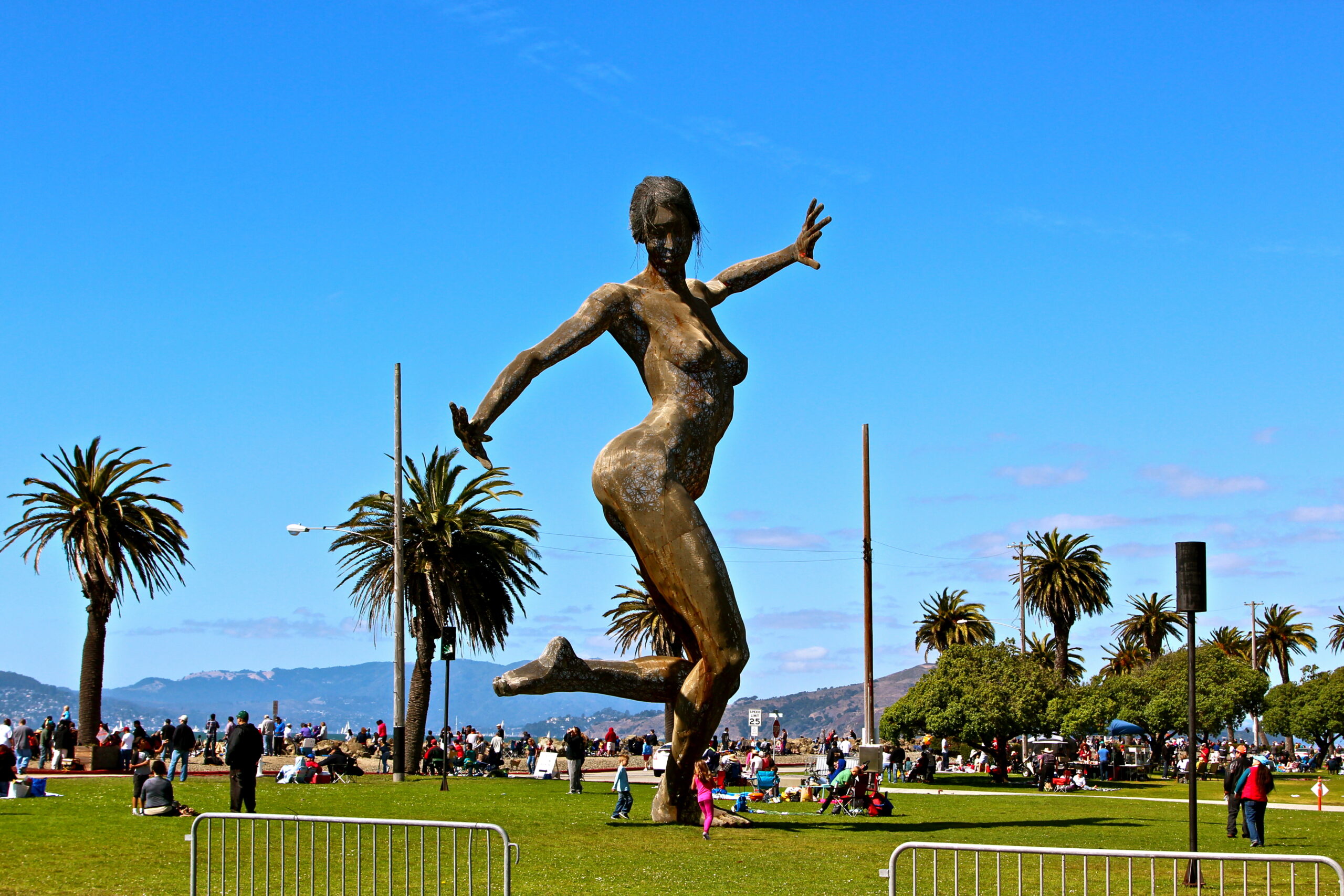 Large outdoor sculpture of a running woman with palm trees and people in the background, sunny day in Lodi, California.