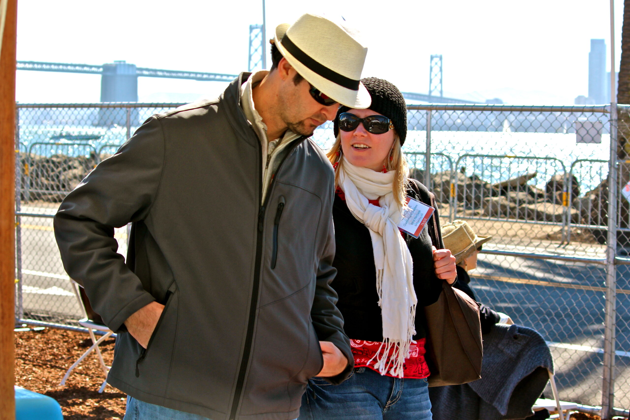 Two people at a Lodi wine event, one with a hat and sunglasses, near a chain-link fence with a bridge in the background.