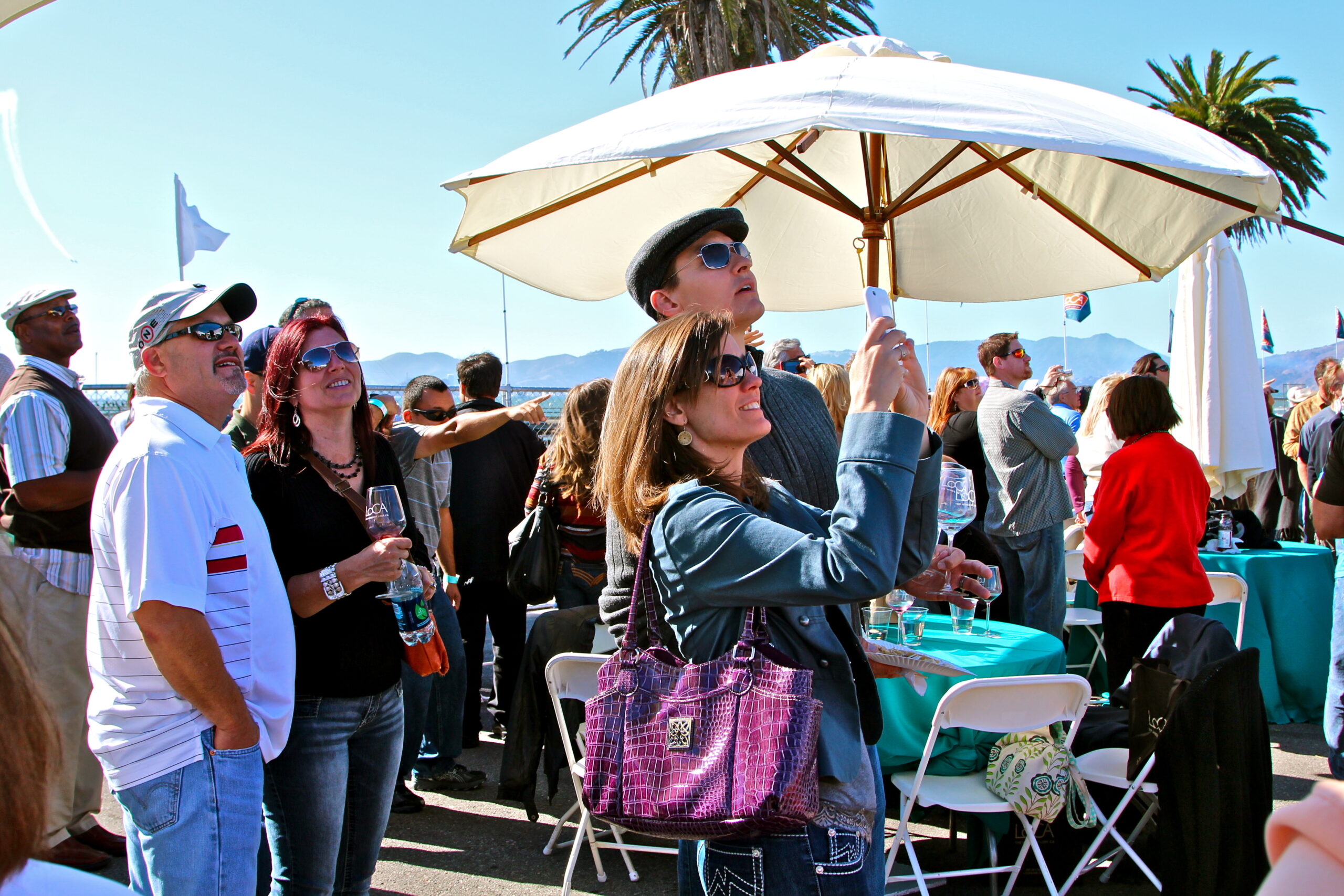 Group of people enjoying a wine event outdoors with a woman holding an umbrella for shade.