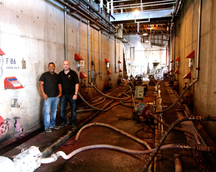 Two men standing inside a wine production facility with pipes and equipment in Lodi, California.