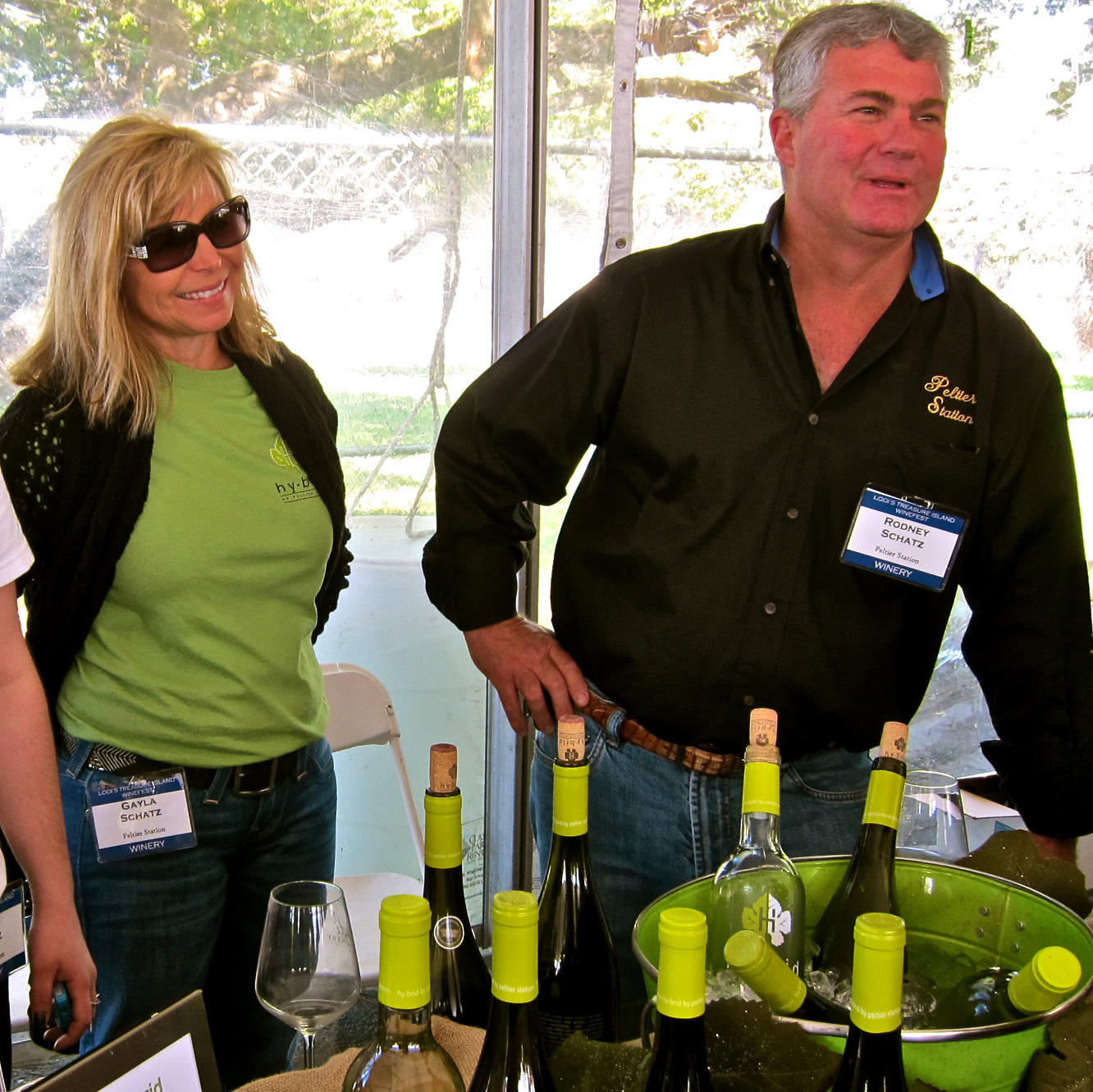 Two people at a wine tasting event with bottles of wine and glasses on a table.